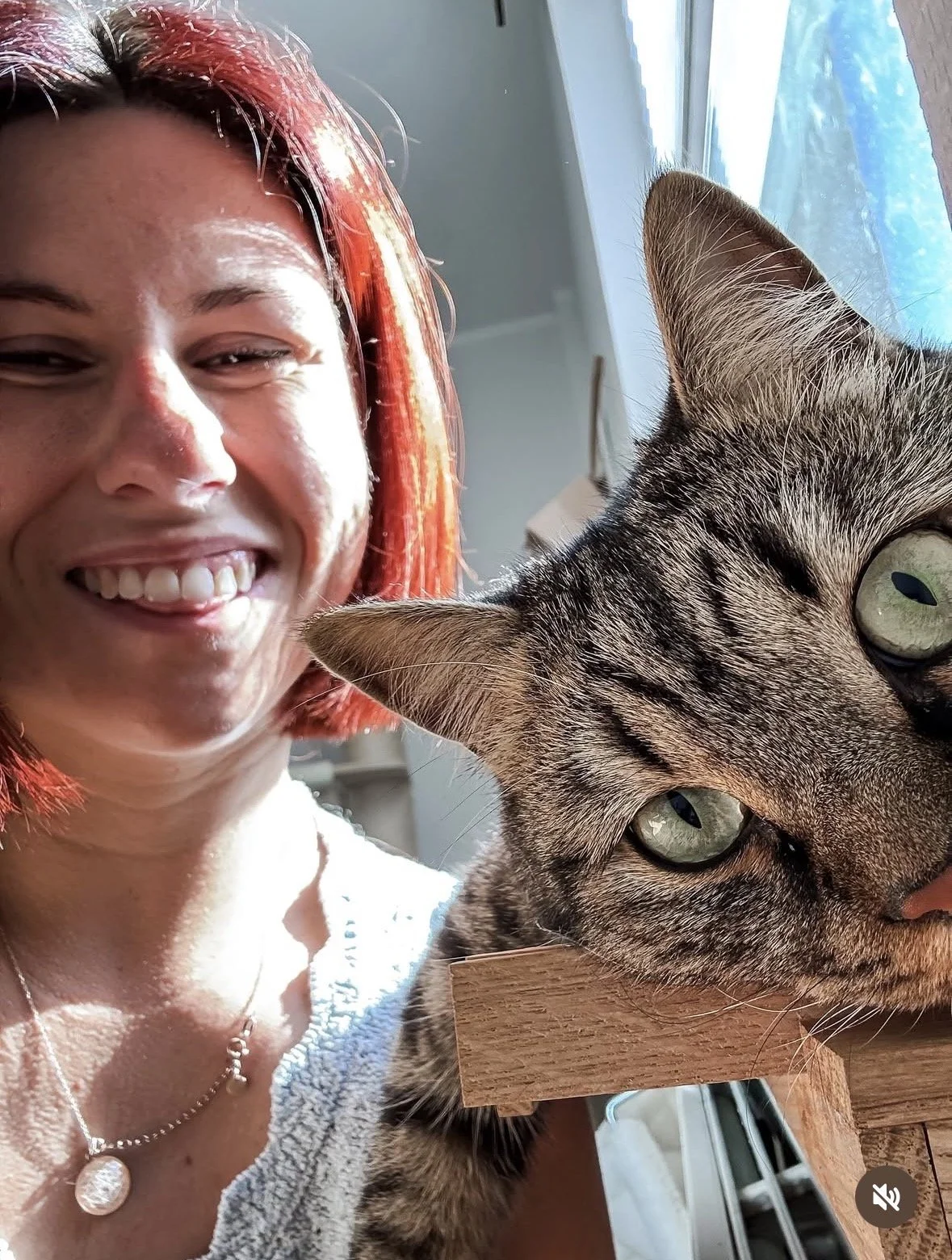 Eine lächelnde Frau mit roten Haaren und eine Katze, die auf einem Holzbrett liegt, neben einem Fenster.