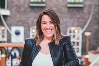 A woman with shoulder-length brown hair smiling, wearing a black leather jacket and white top, standing outdoors with a brick building and string lights in the background.