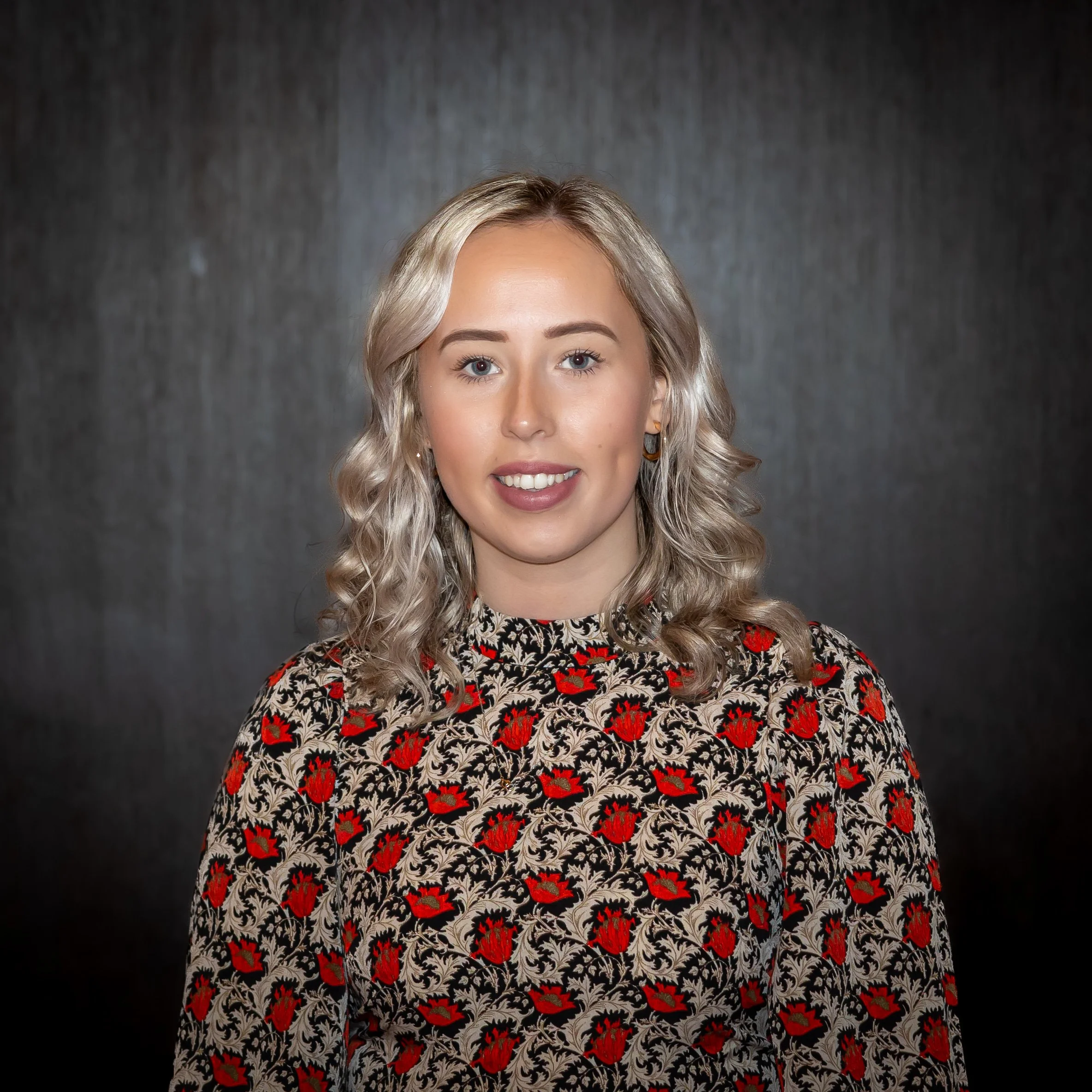 A young woman with blonde hair and blue eyes standing outdoors in a park with trees and grass, wearing a black vest with an embroidered patch that reads 'Floristry' and a gray long sleeve shirt underneath.