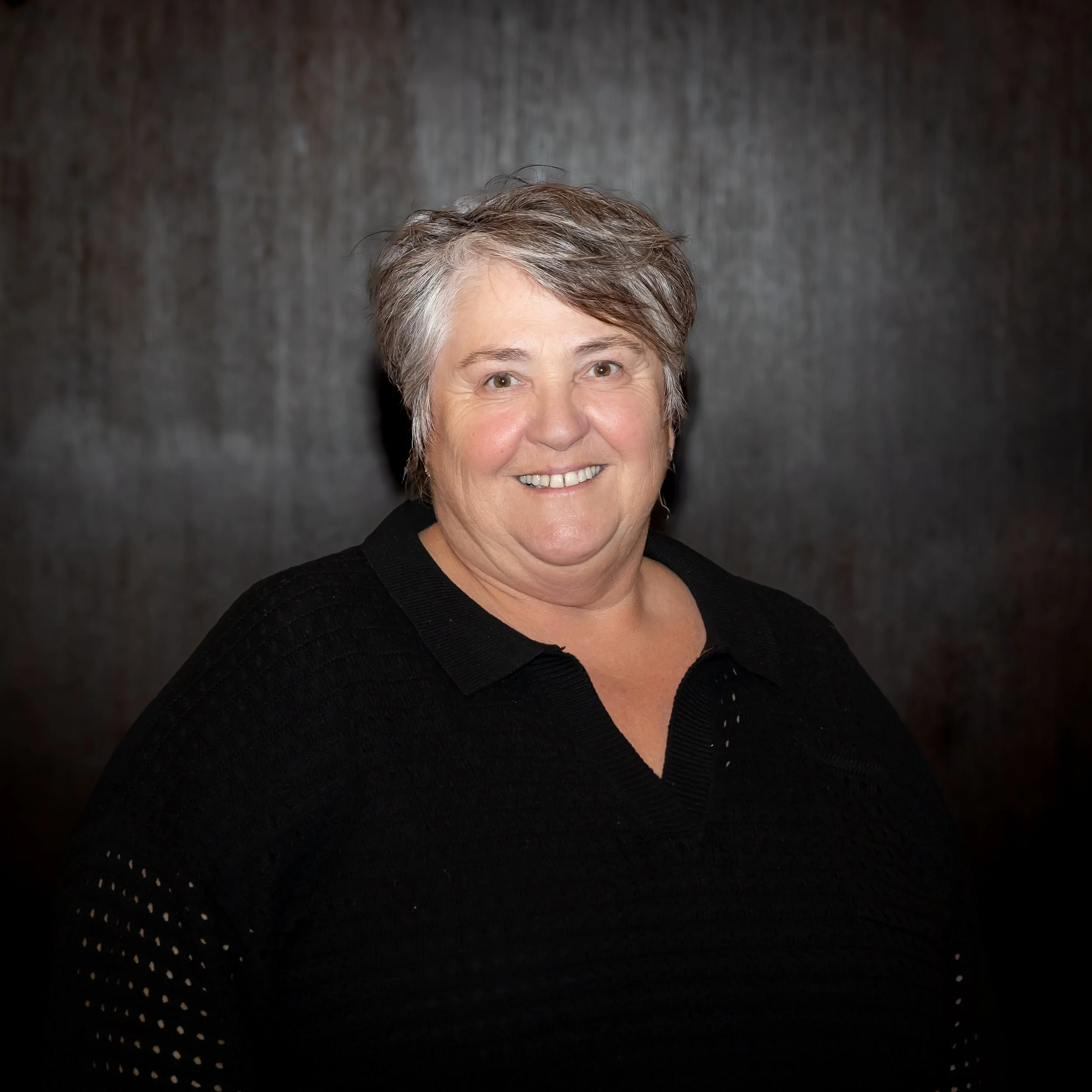 A smiling middle-aged woman with short, salt-and-pepper hair, wearing a black lace top, posing outdoors against a stone wall.