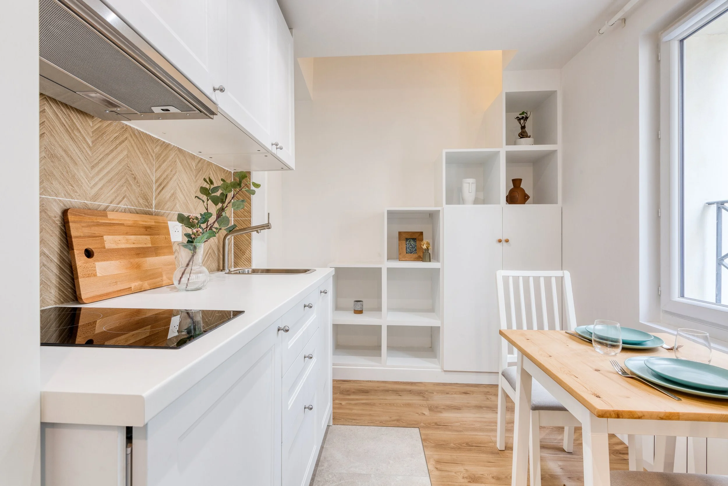 Cuisine moderne avec comptoir blanc, poêle, planche à découper, vase avec feuillage, étagères blanches, table et chaises en bois avec assiettes vertes, murs et sol en bois clair.