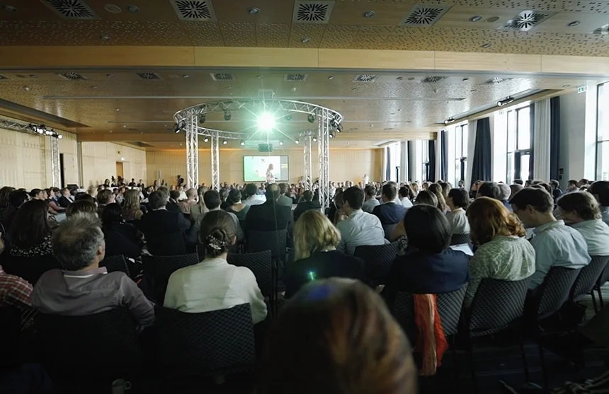 Ein großes Publikum, das in einem Konferenzraum sitzt und eine Präsentation auf einer Bühne mit einem Bildschirm und heller Beleuchtung verfolgt.