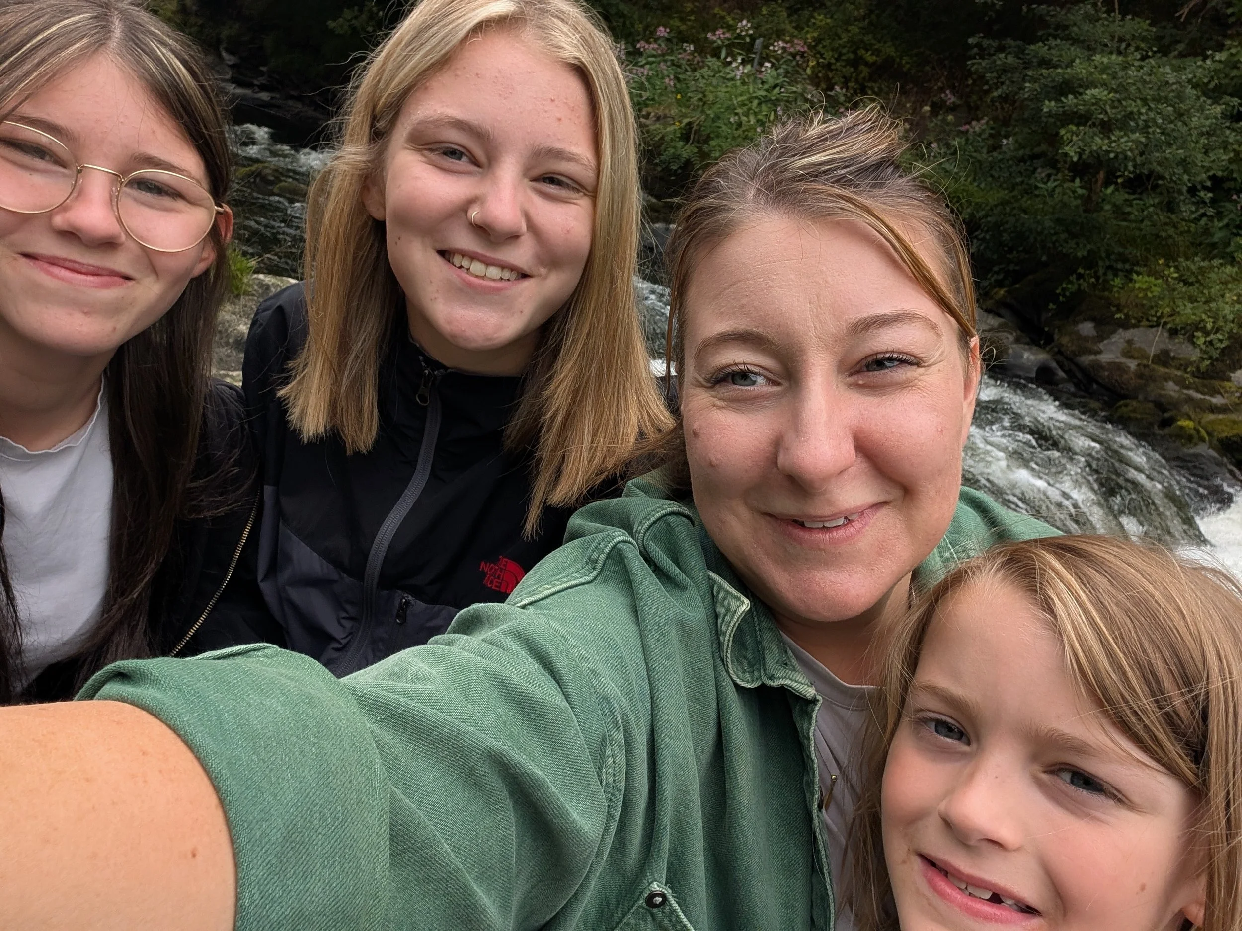 A group of five people taking a selfie outdoors near a flowing creek, with trees and greenery in the background.