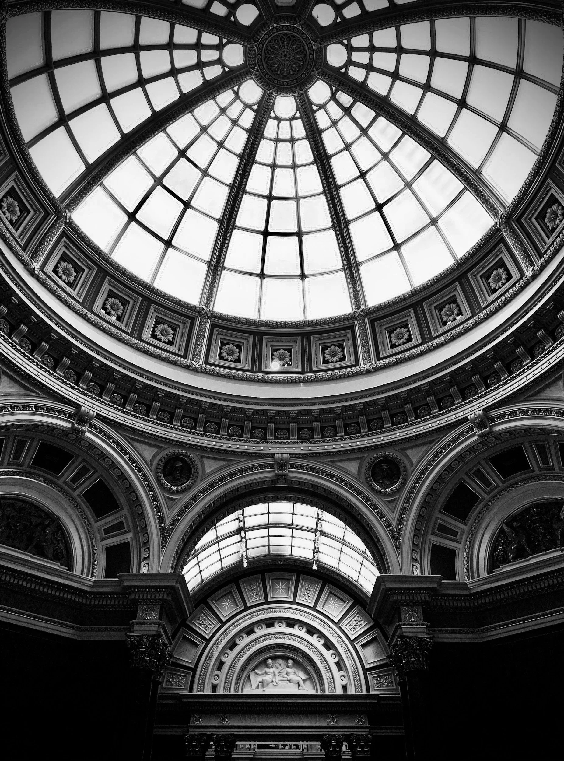 The interior of a historic building with a large glass dome ceiling, ornate architectural details, and decorative sculptures, photographed in black and white.
