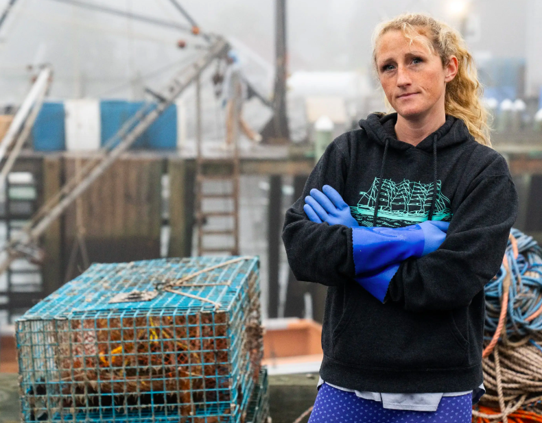 A person with curly hair standing with arms crossed, wearing a black hoodie and blue gloves, beside a lobster trap on a dock.
