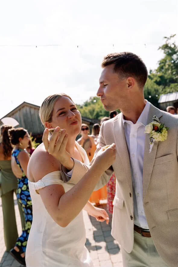 A man in a suit and a woman in a white dress eat oysters at an outdoor event with trees in the background.