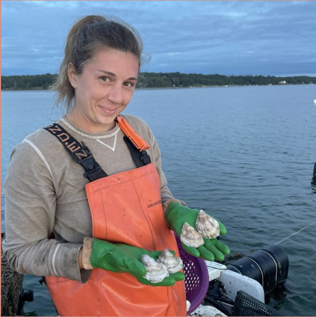 Person in orange overalls holding clams by the water