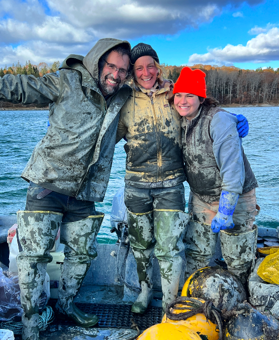 Three people wearing muddy overalls and boots standing on a boat, smiling and posing for a photo with a backdrop of water and trees, one person wearing a red beanie.