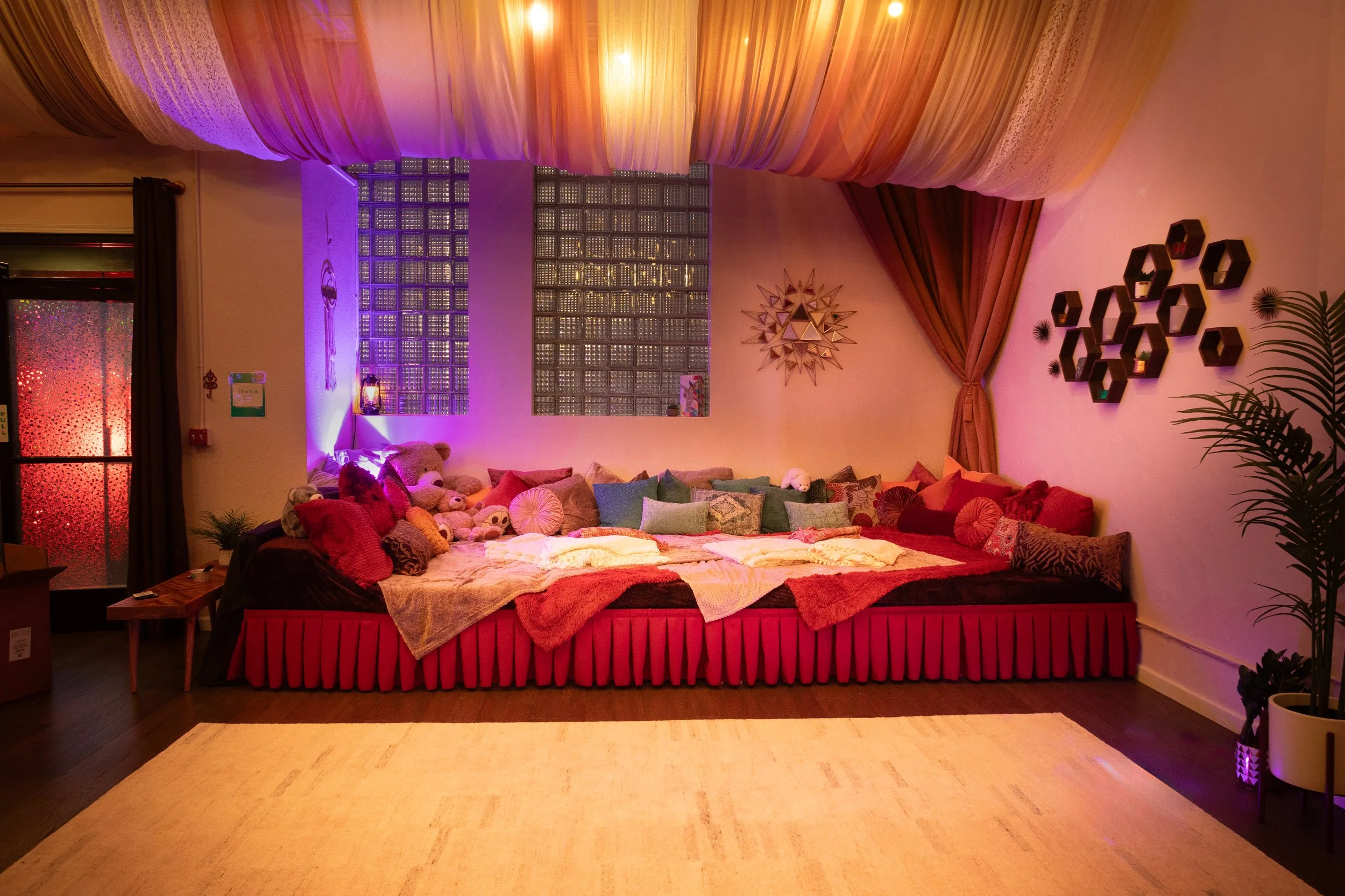 A cozy corner decorated with plush toys, cushions, and soft lighting, featuring a large bed or lounging area, colorful pillows, wall decor, and purple ambient lighting.