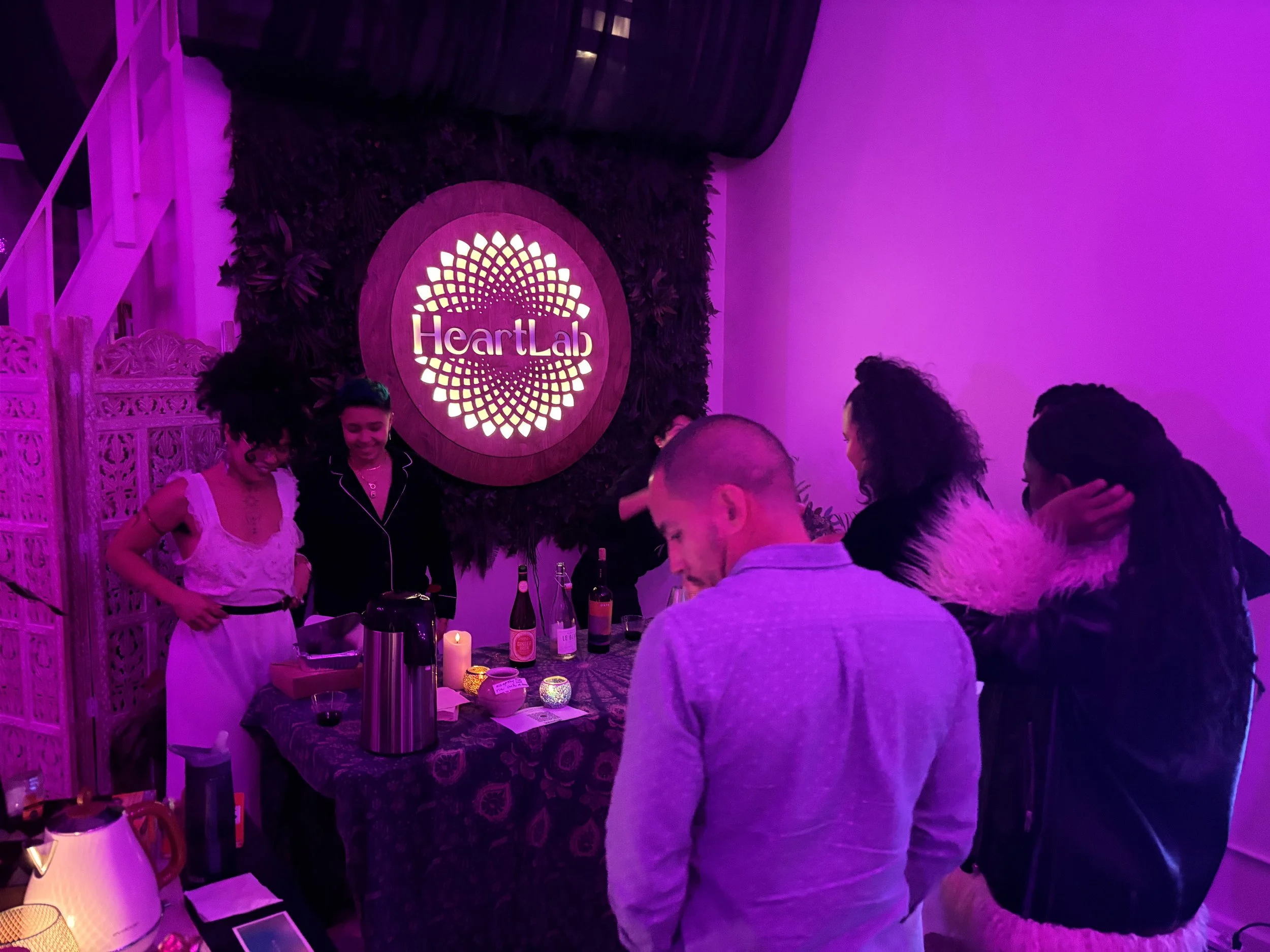 Group of people gathered around a table at Heart Lab event, illuminated by pink/purple lighting, with drinks and candles on the table, and a Heart Lab sign on the wall.