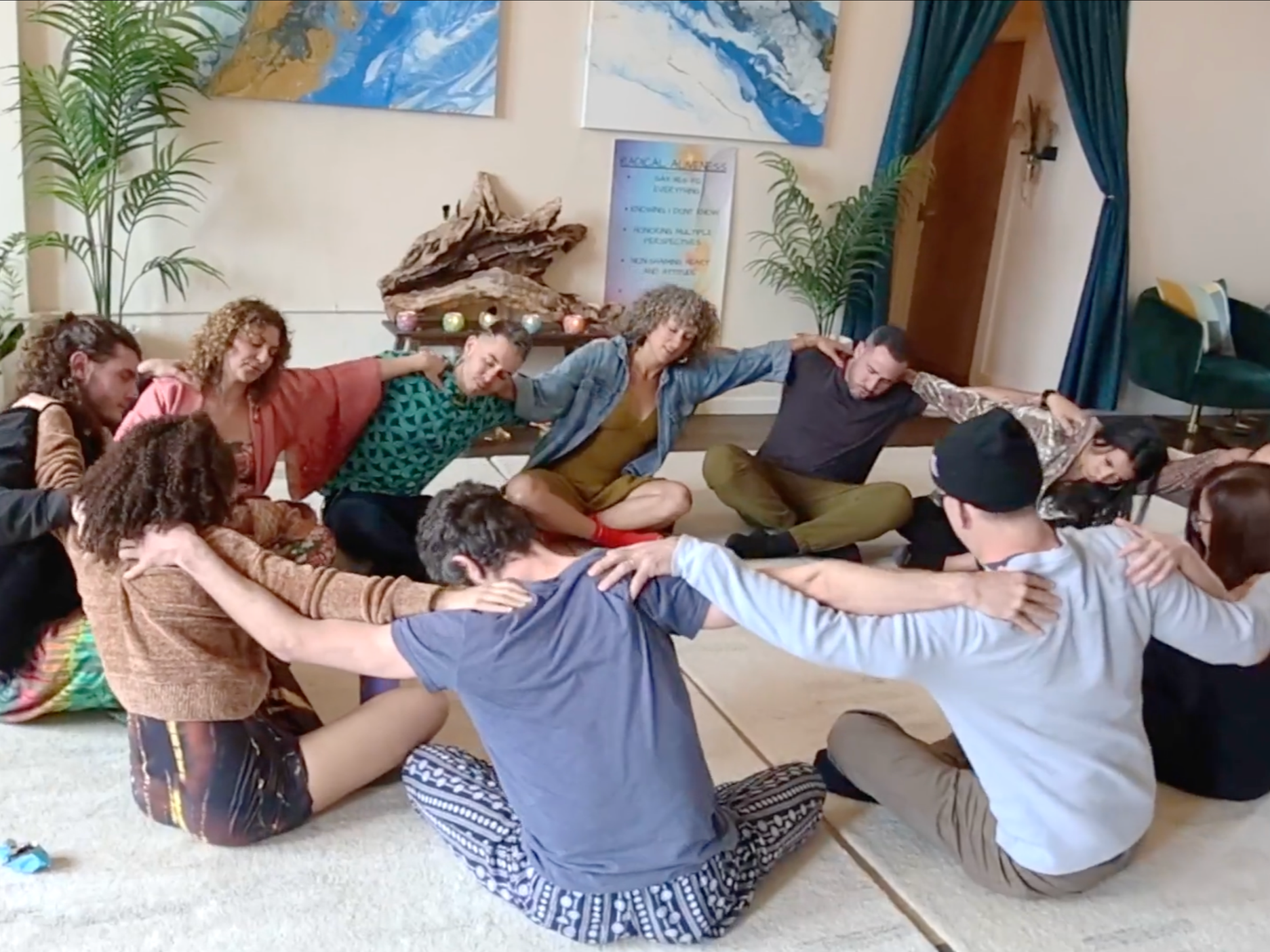 A group of people sitting on the floor in a circle, holding each other’s shoulders, participating in a group activity or meditation in a cozy indoor setting.