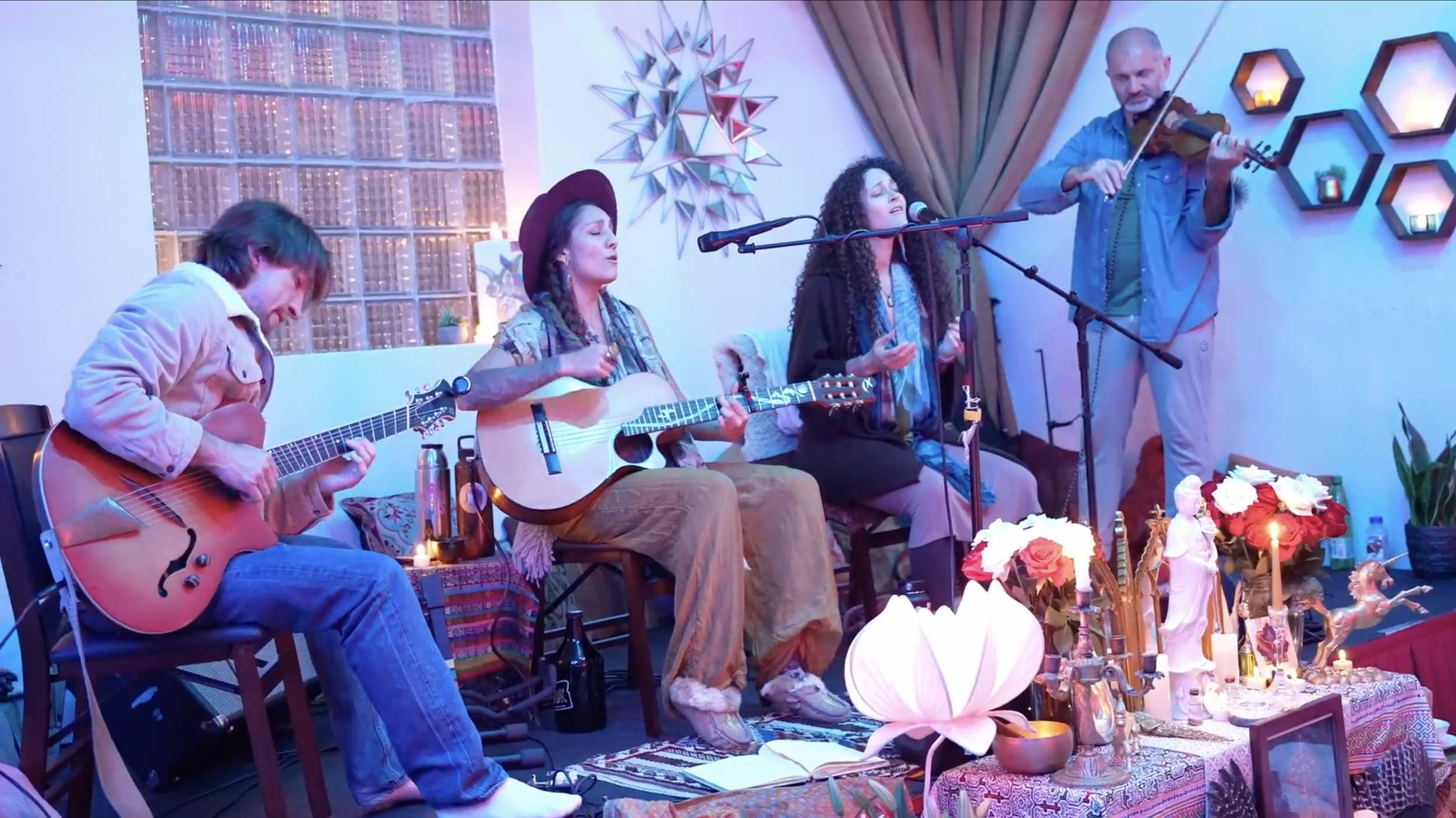 Four musicians performing indoors, three women and one man, with guitars and a violin, with decorative elements and candles on a table in front.