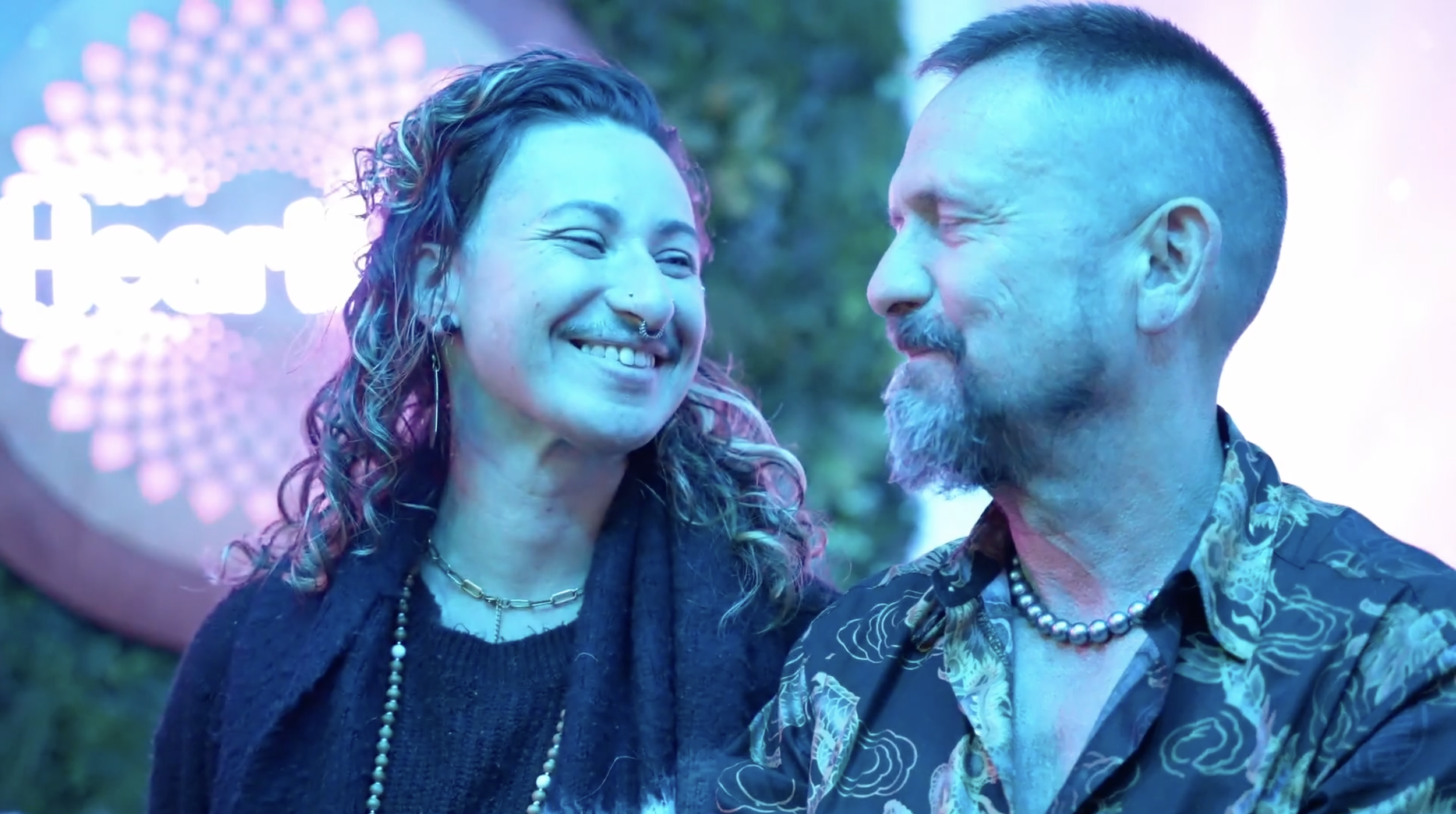 A woman and a man smiling at each other at an event with colorful lighting and a sign that says 'heart' in the background.