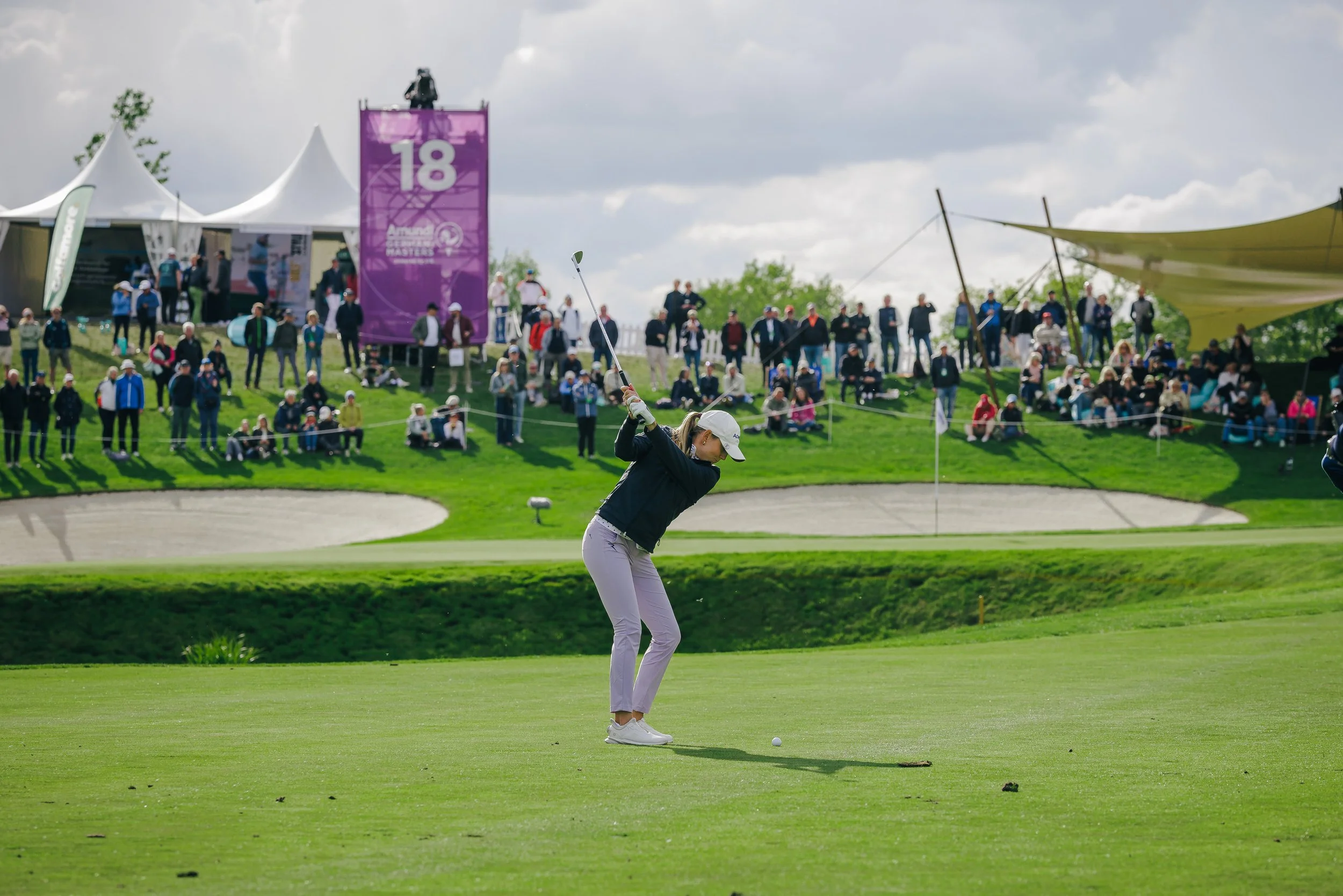 A female golfer teeing off on a golf course as spectators watch from the hillside behind her during a tournament.