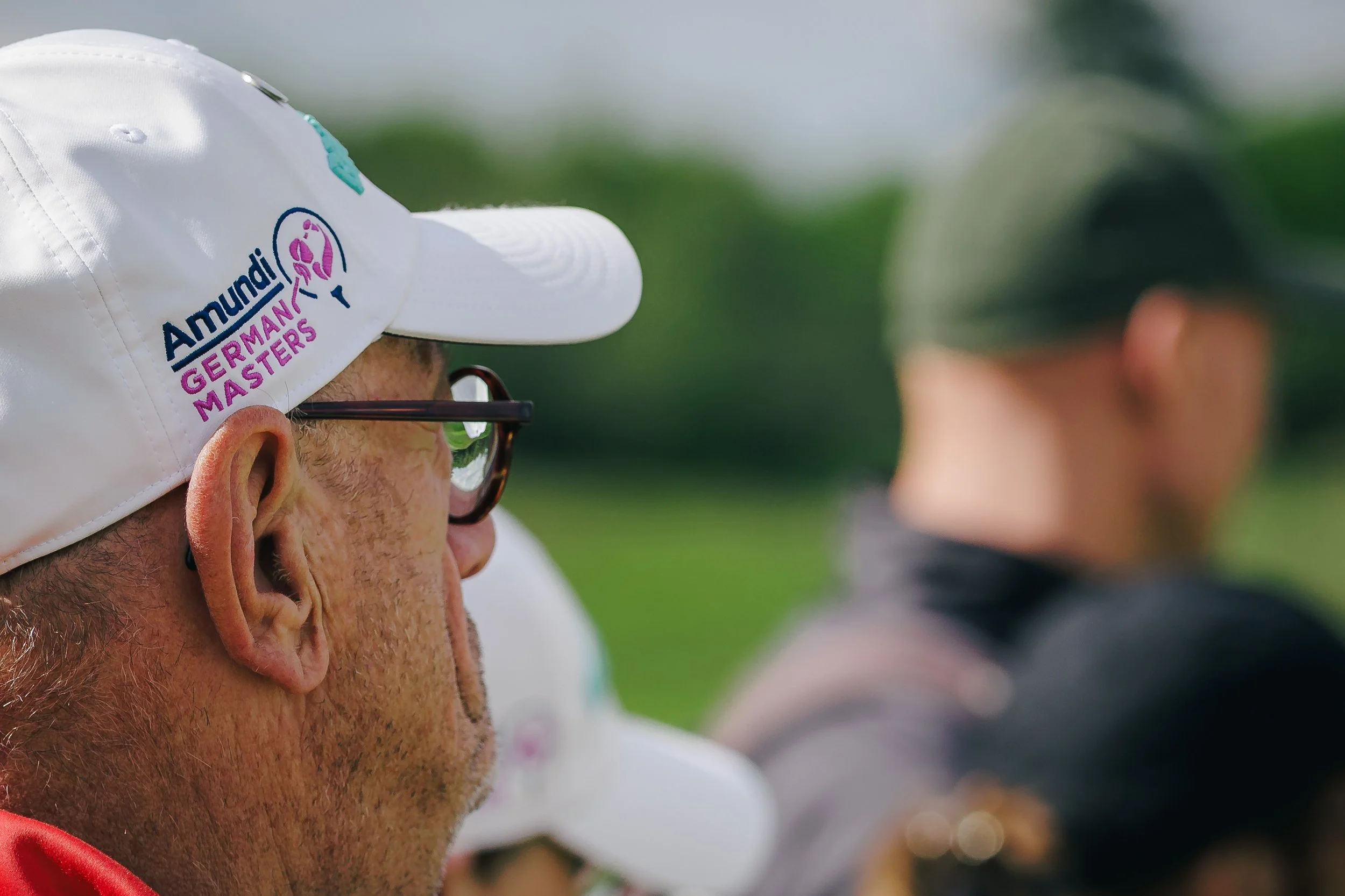 Close-up of a man wearing a white cap with logos and glasses. The background shows a blurred outdoor setting with greenery and people.