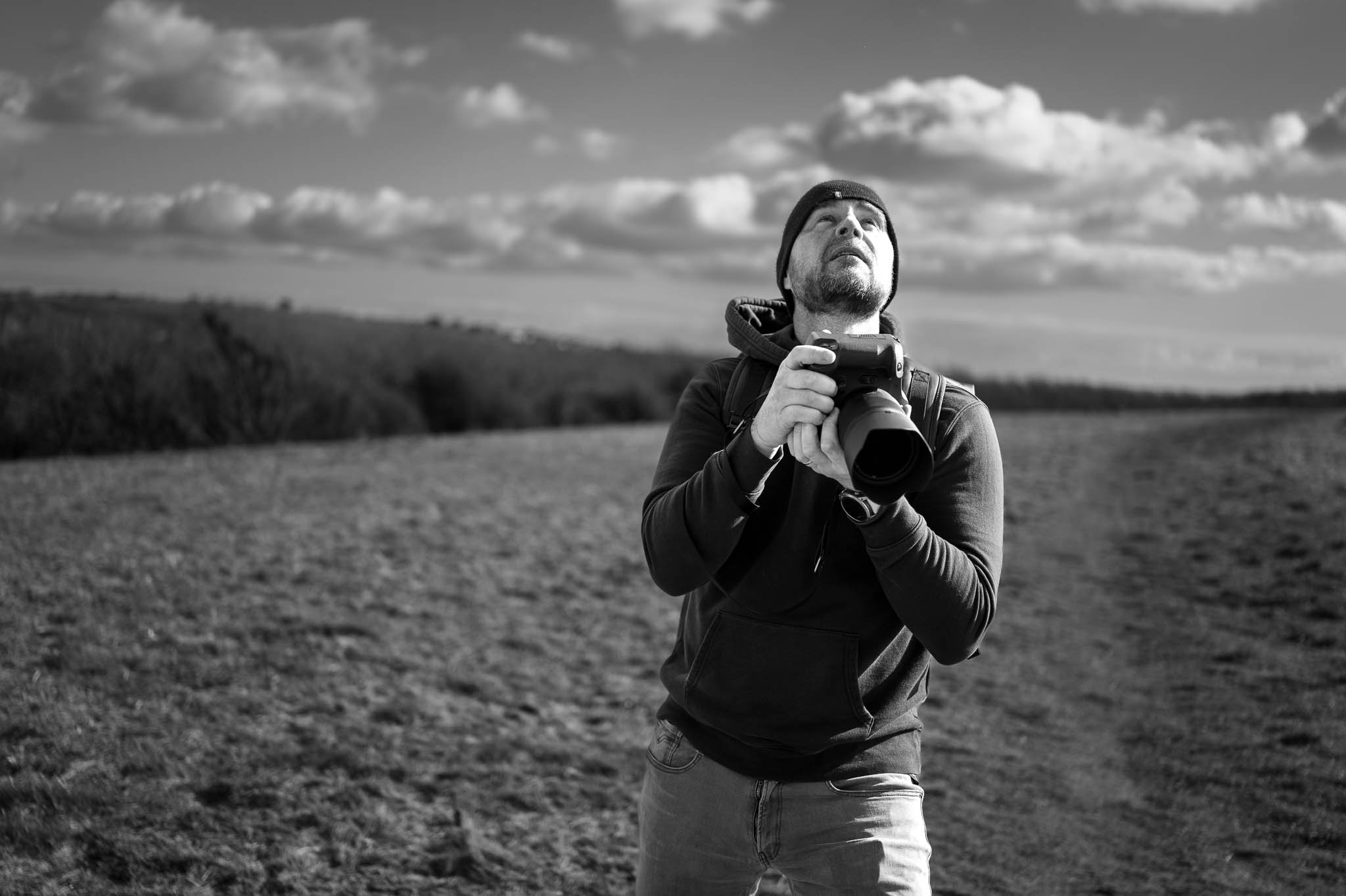 Black and white photo of a man outdoors holding a camera and looking upwards, with a landscape and cloudy sky in the background.