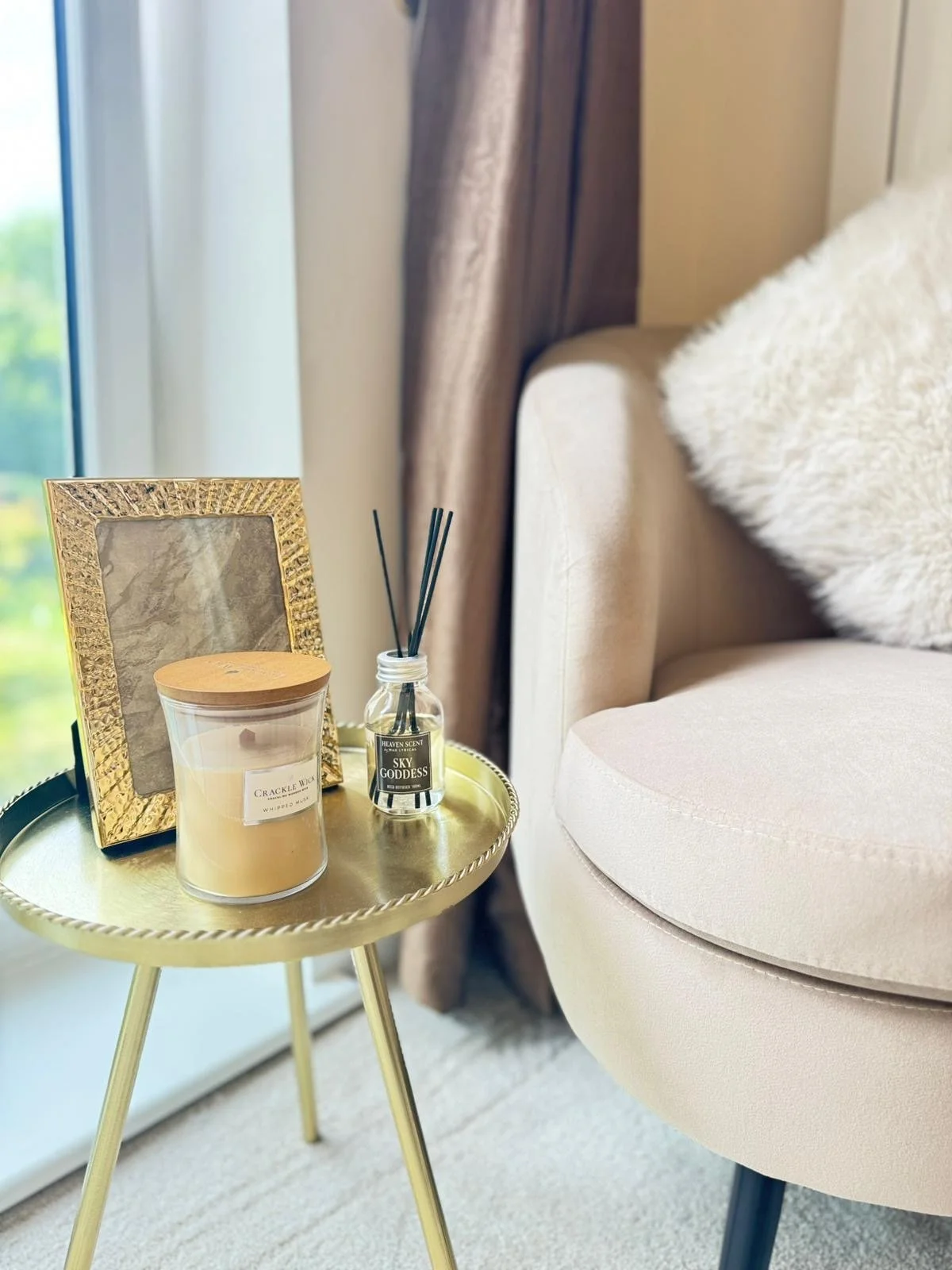 A small gold side table next to a beige armchair with a white fluffy pillow. The table holds a gold picture frame, a scented candle, and a reed diffuser.