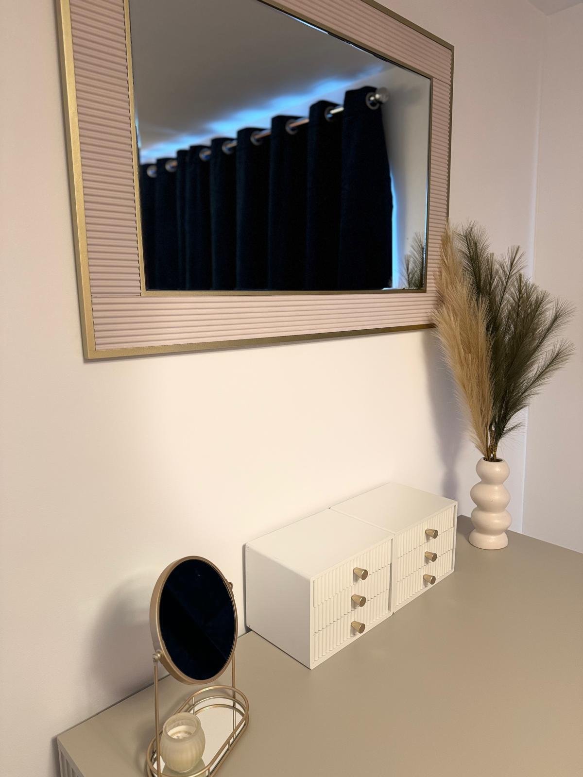 A decorative setup on a light-colored table featuring a small round mirror with a stand, a white box with gold knobs, and a tall white vase with dried pampas grass. A large rectangular mirror with a textured beige frame hangs on the wall above, refle
