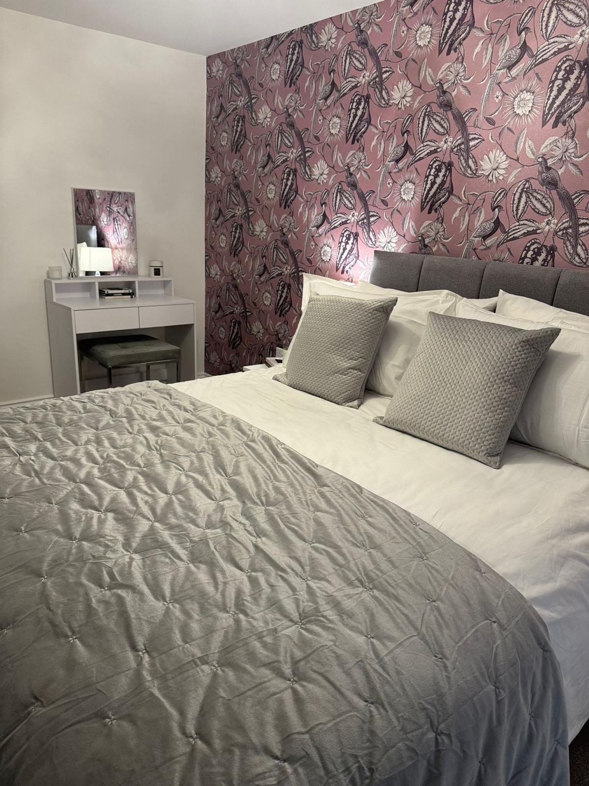 A neatly made bed with a gray quilted bedspread, four pillows (two gray and two white), against a gray upholstered headboard. Behind the bed is a pink accent wall with a floral and bird pattern. To the left, a small white vanity with a mirror, a lamp, a reed diffuser, and a few items on it.