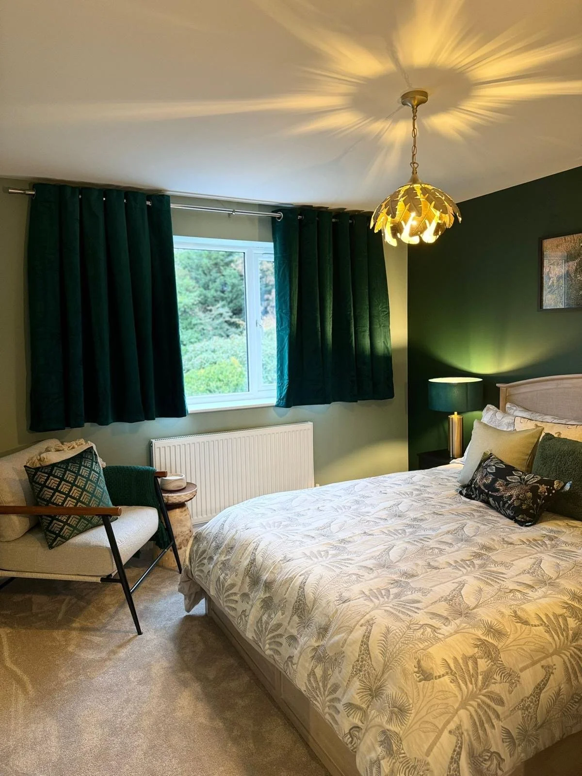 A bedroom with green curtains, a window showing trees outside, a bed with tropical-patterned bedding, a nightstand with a lamp, a framed picture on the green wall, a beige headboard, and an armchair with pillows near the window.