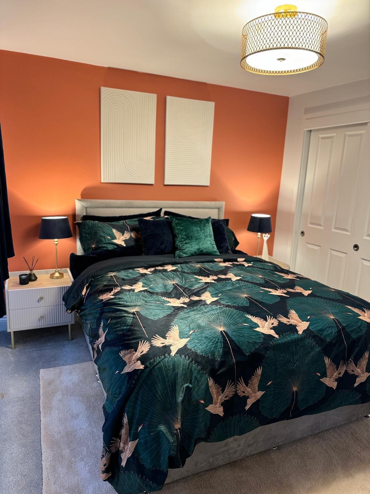 A bedroom with a bed featuring dark green and beige bedding with bird and leaf patterns, two matching black nightstands with black lamps, a coral accent wall with two abstract white art pieces, a white closet with sliding doors, a modern ceiling ligh
