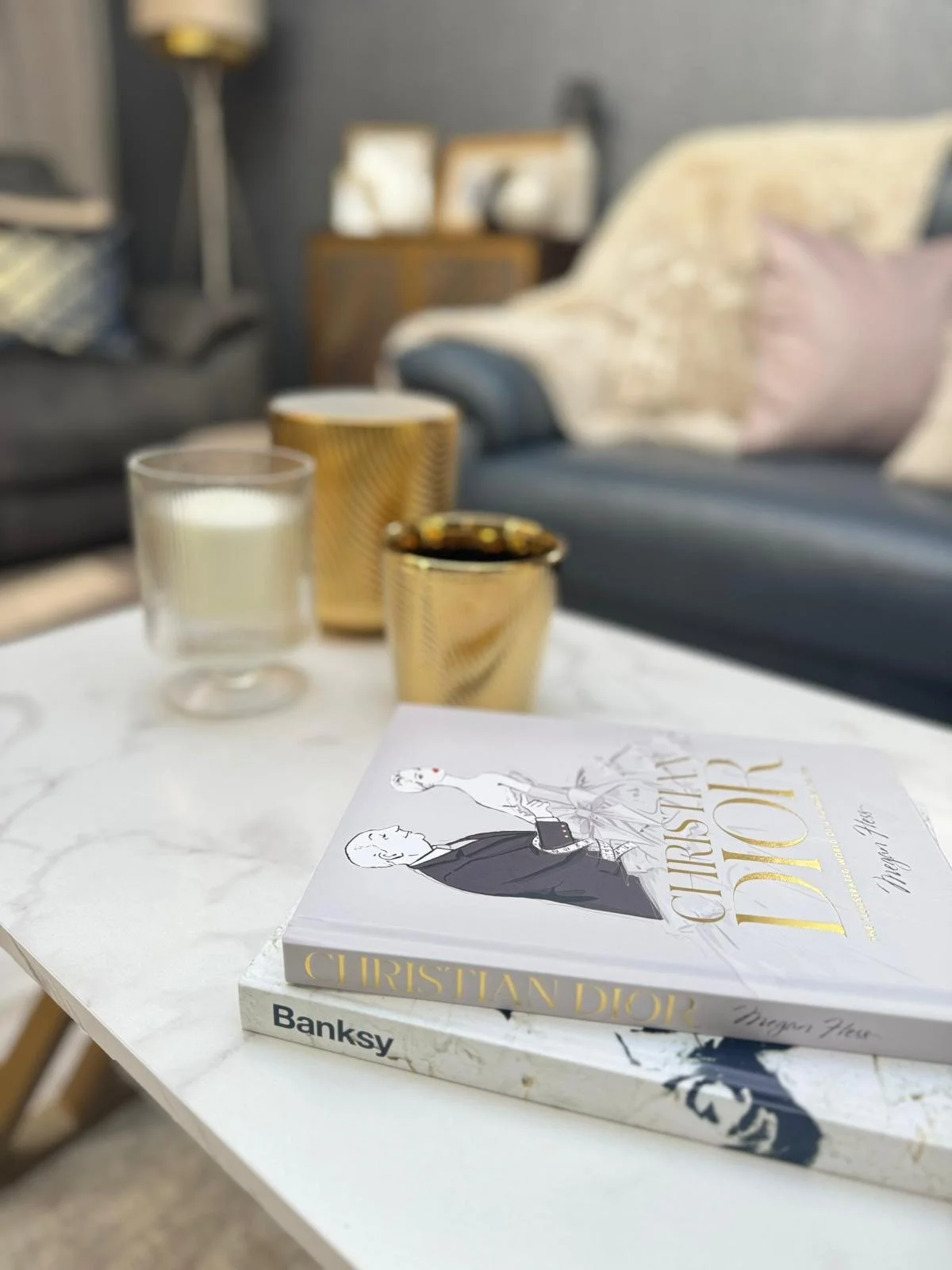 A white marble coffee table with two books, one titled "Christian Dior" and the other "Banksy." On the table, there are also three candles in gold and clear holders, and a glass candle holder with a white candle inside.