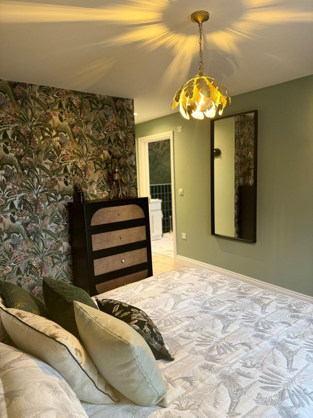 A bedroom with a tropical-themed accent wall, a bed with cream and black pillows, a mirror on the light green wall, a black headboard, and a decorative ceiling light casting shadows on the ceiling.