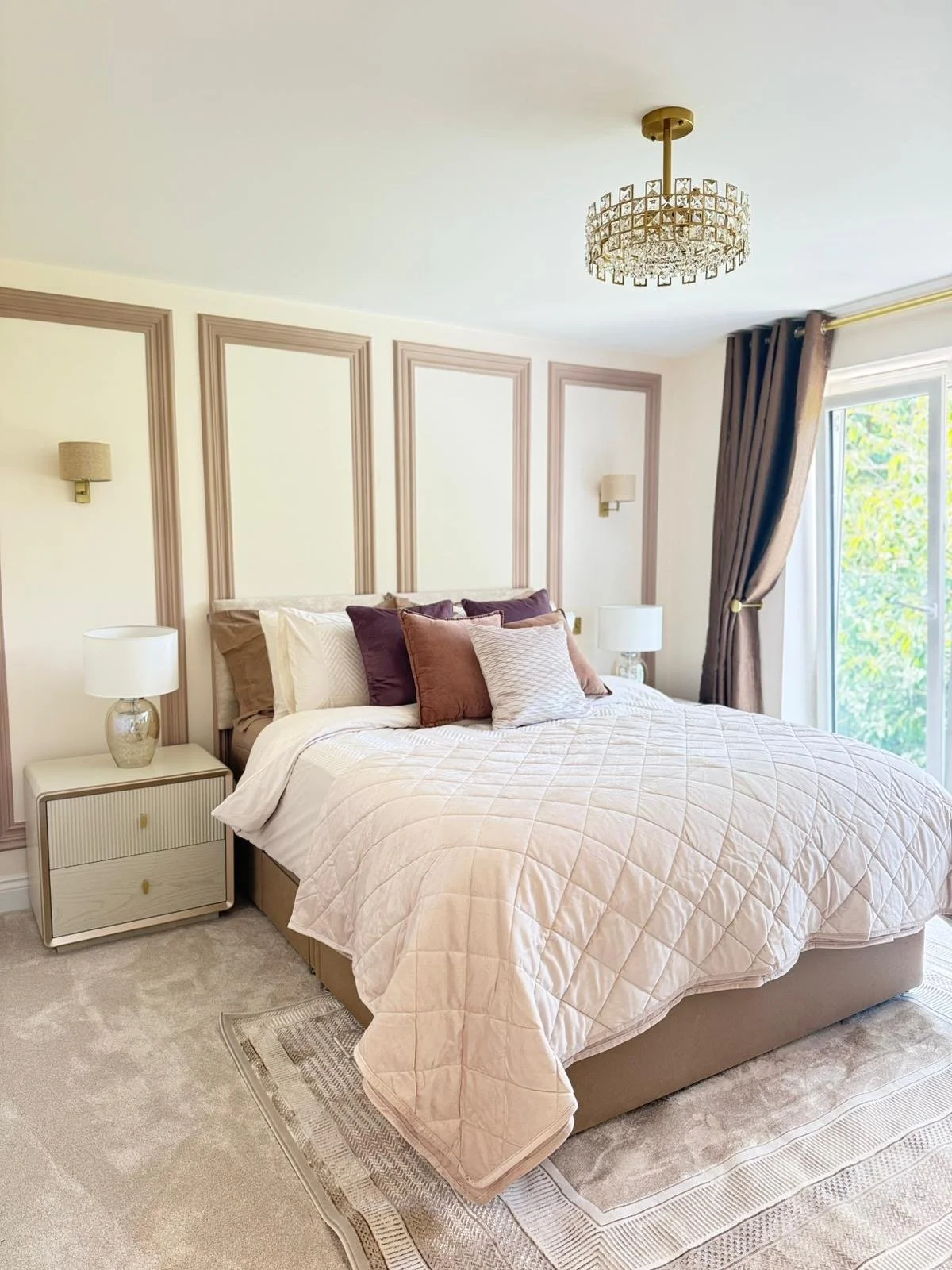 A neatly made bed with multiple pillows in shades of purple, brown, and beige, in a bedroom with cream-colored walls, decorative wall sconces, a chandelier, side tables with lamps, beige curtains, and a sliding glass door with greenery outside.