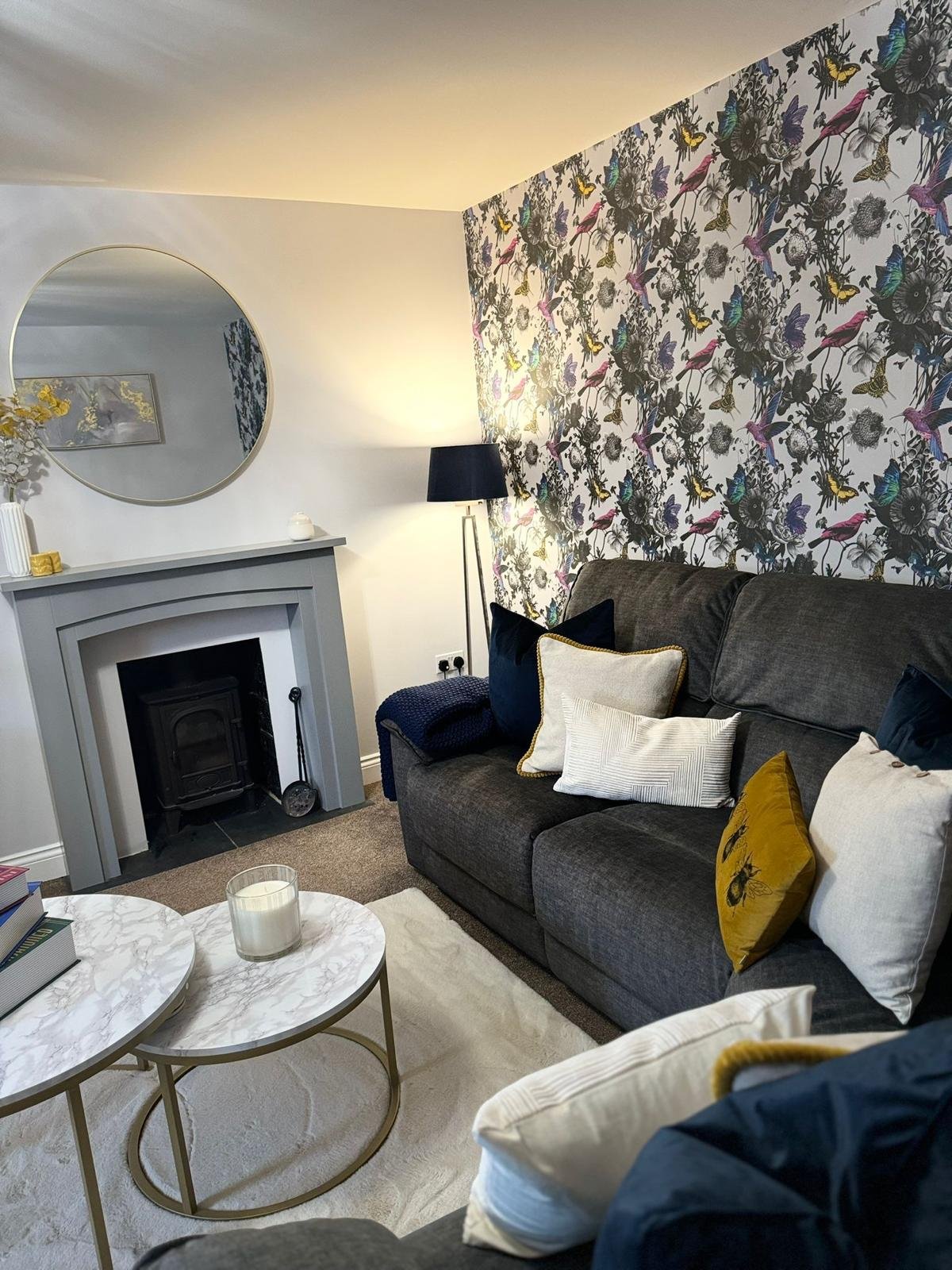A cozy living room with a dark gray sofa adorned with various pillows, a white rug, two overlapping marble coffee tables with a candle, a fireplace with a mirror above, a black floor lamp, a feature wall with vibrant butterfly and bird patterned wall