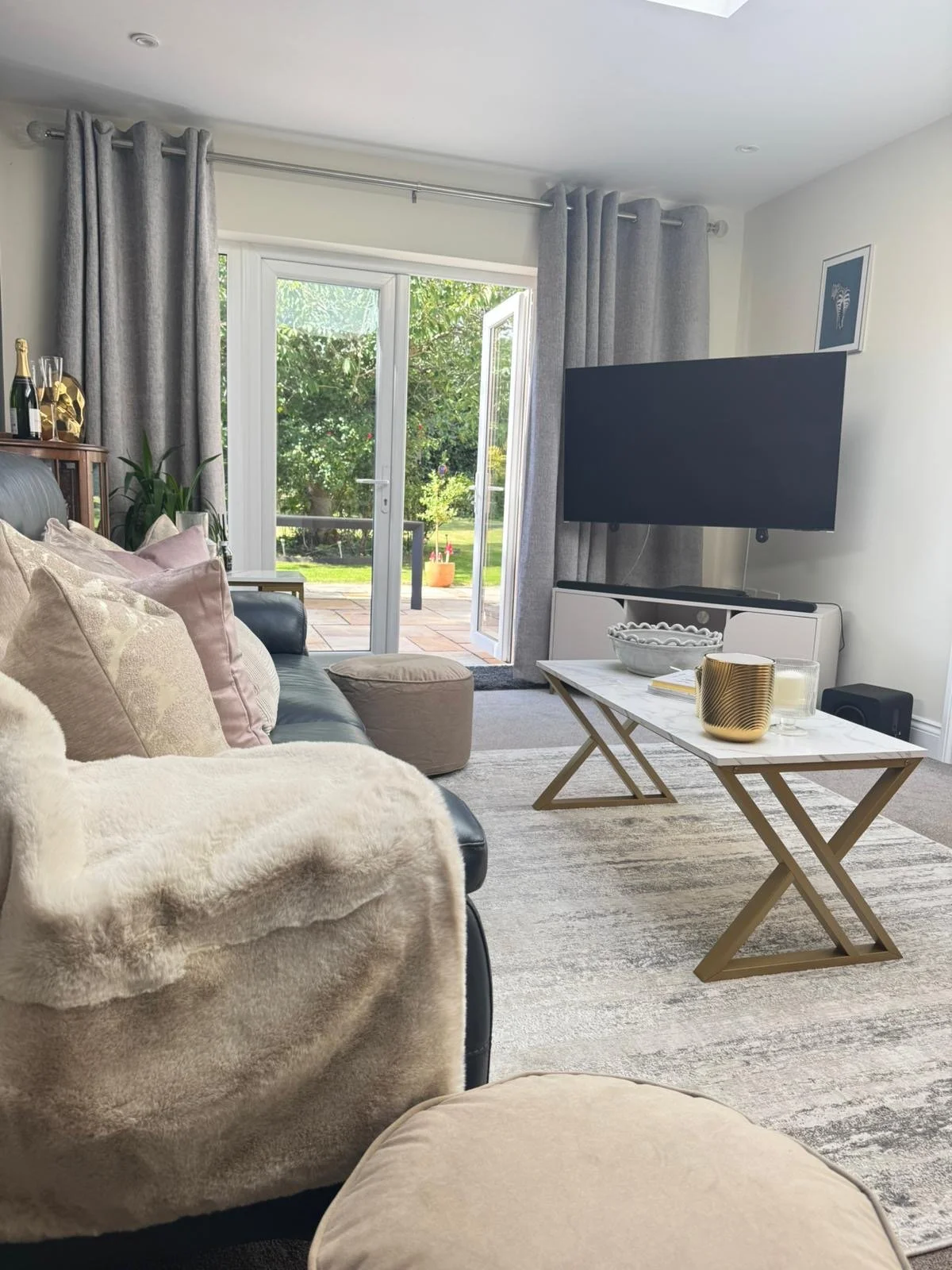 Living room with a sofa, plush pillows, a coffee table with decorative items, and a TV, with an open door leading to a patio and garden.