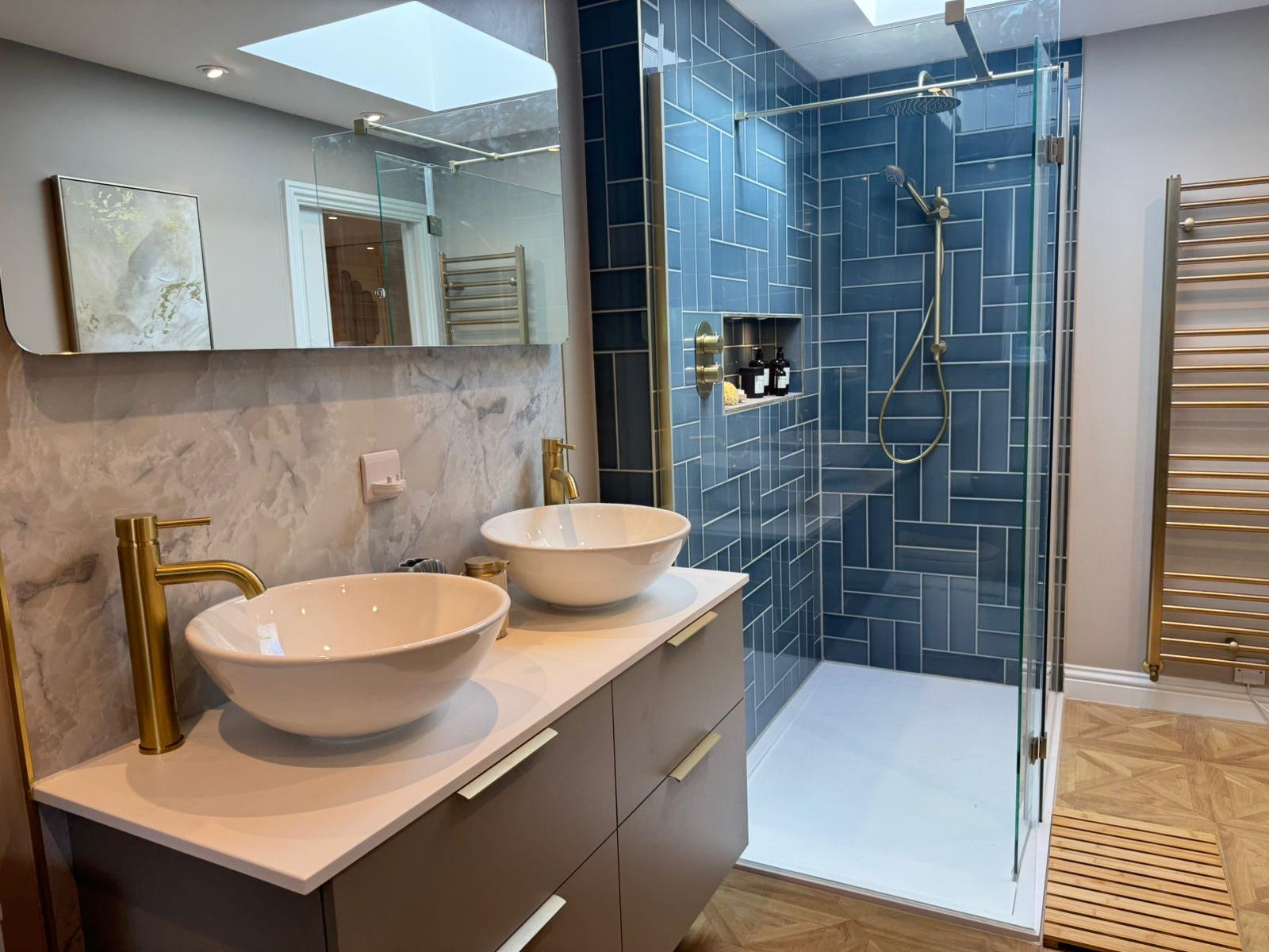 Modern bathroom with a double vanity featuring two vessel sinks with gold fixtures, a marble backsplash, and a large mirror. There is a separate glass-enclosed shower with blue tiles and built-in storage. The bathroom has wood flooring and a heated t