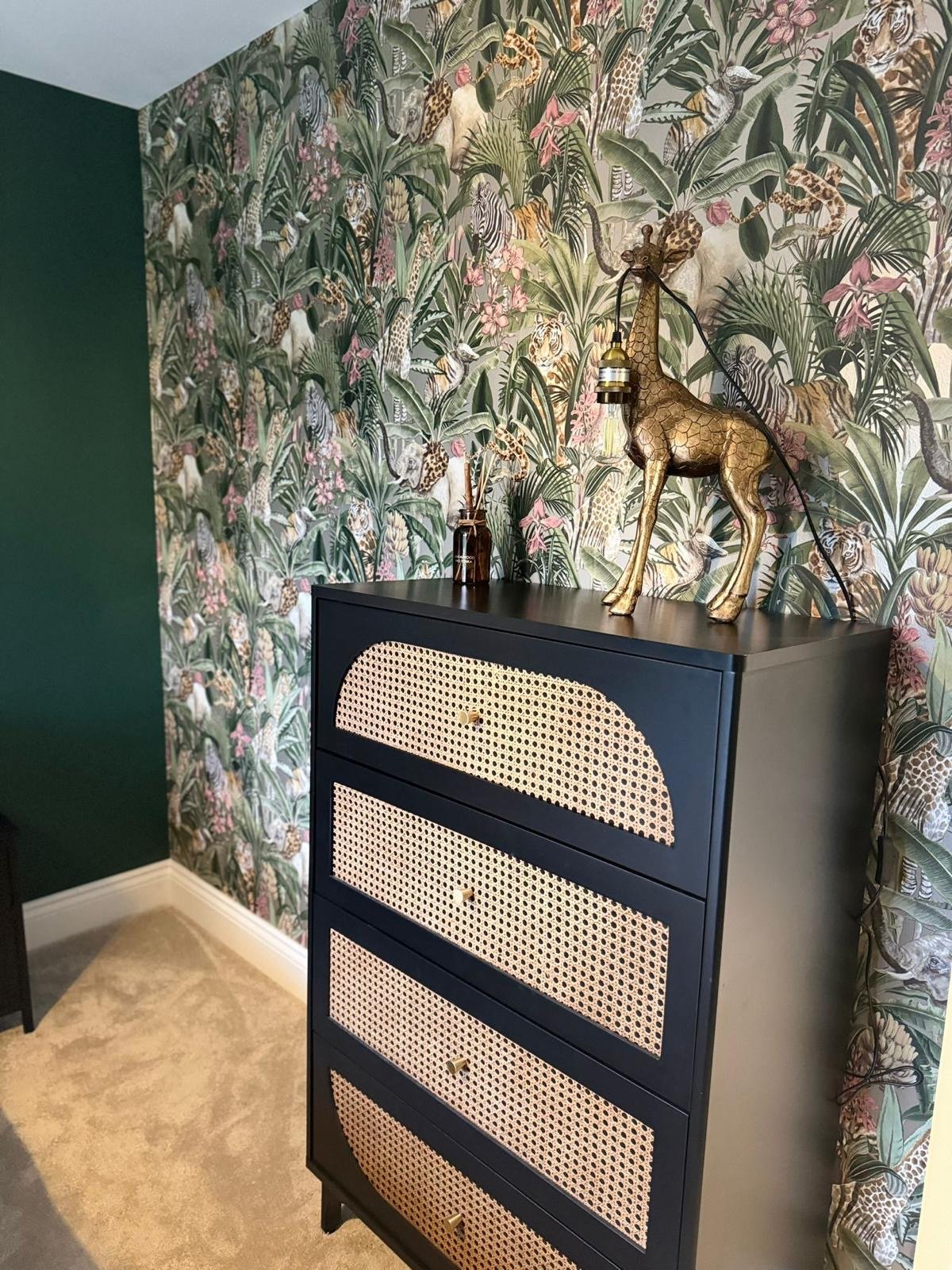 A black dresser with cane-front drawers in a room with tropical-themed wallpaper featuring animals such as tigers, leopards, and giraffes, decorated with a gold giraffe sculpture, a small vase, and a hanging lamp.