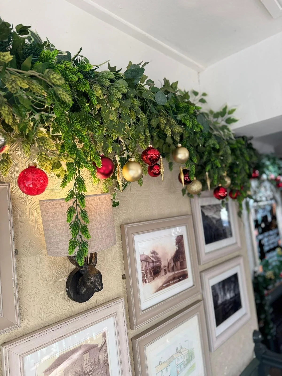Green garland with red, gold, and silver Christmas ornaments hanging on a wall above framed pictures.