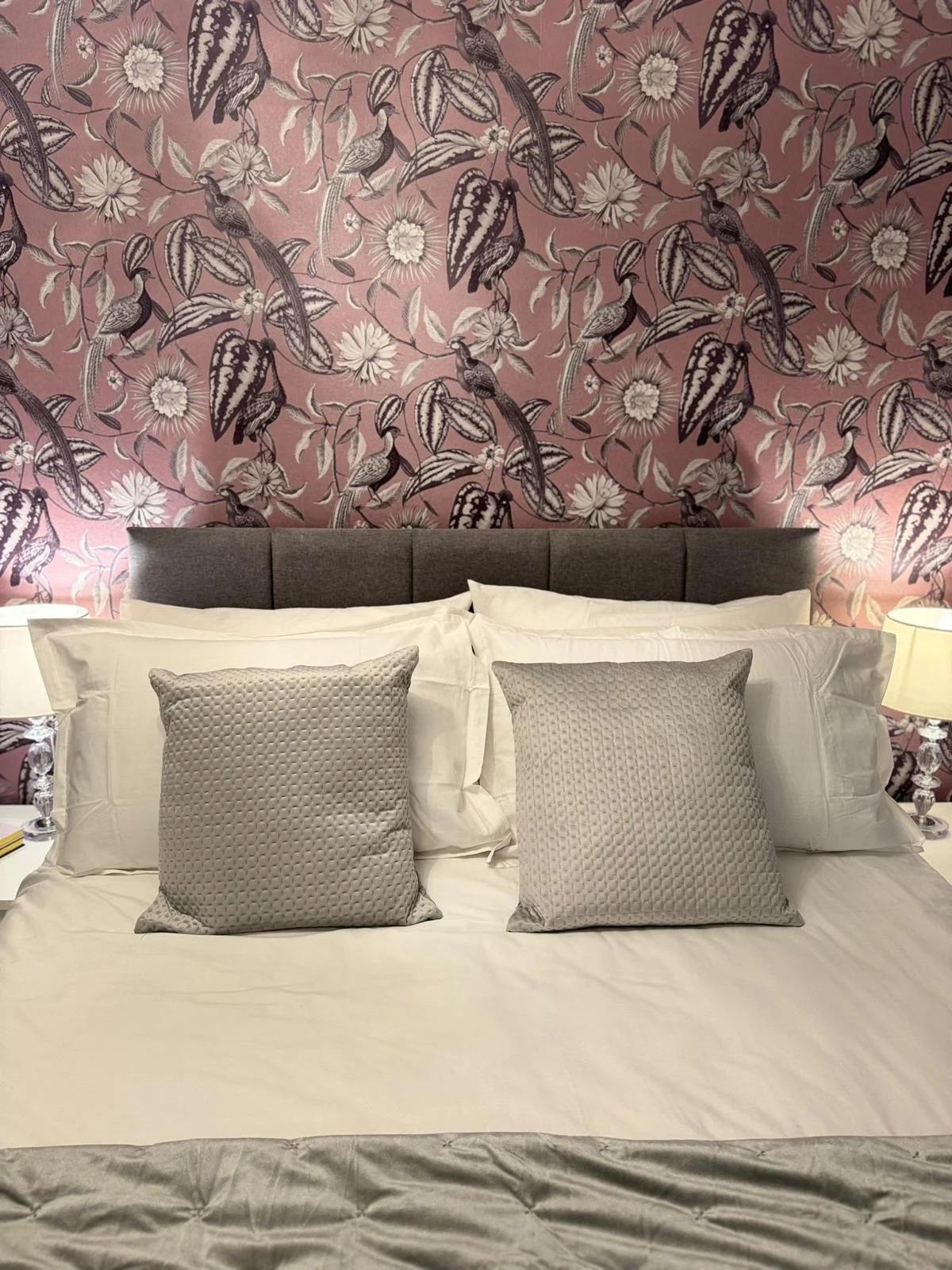 A neatly made bed with a grey upholstered headboard, white sheets, and beige textured accent pillows. Two bedside lamps with white shades are seen on either side of the bed, and the wall behind features pink floral and bird wallpaper.