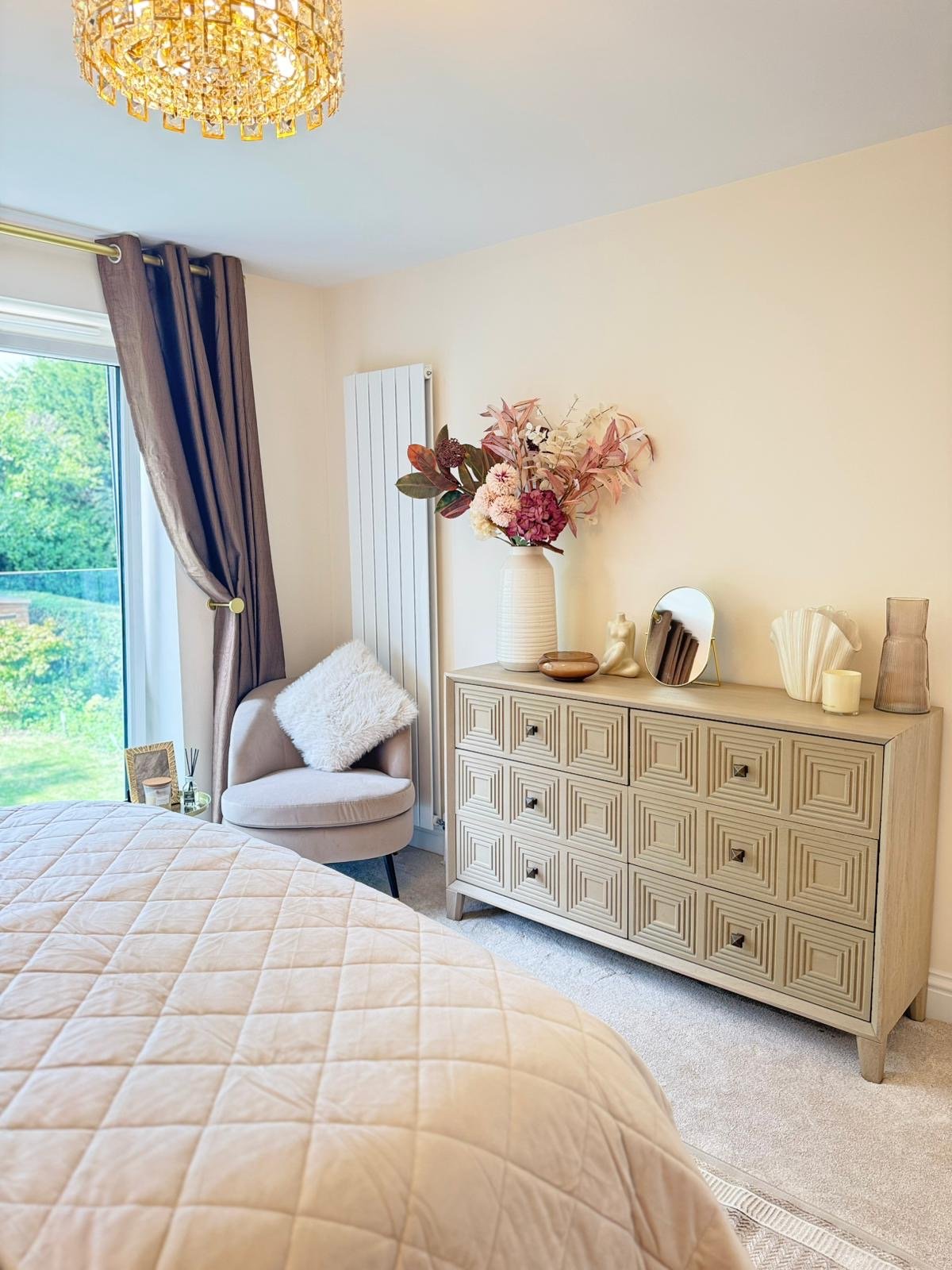 A cozy bedroom corner featuring a beige quilted bedspread, a soft pink upholstered chair with a fluffy white pillow, a light wood dresser decorated with vases, a mirror, candles, and a large floral arrangement, with a window draped in purple curtains