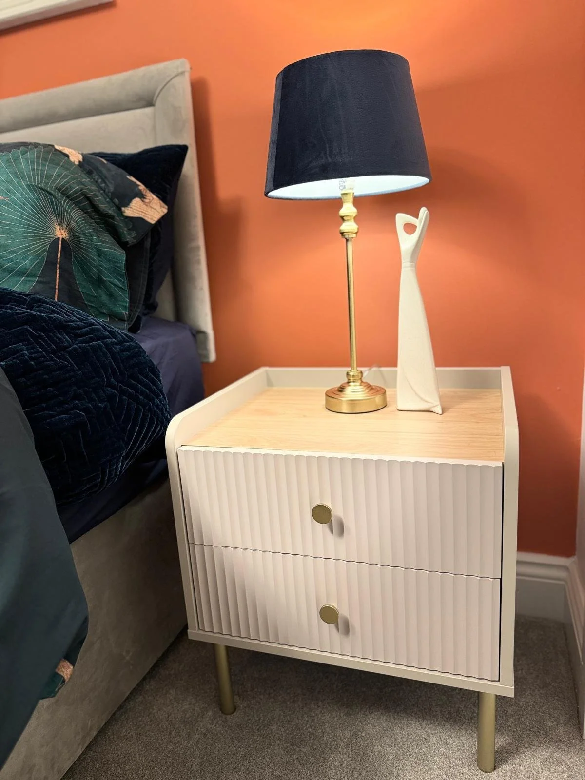 A white bedside table with two drawers and round brass knobs. On top of the table is a gold-colored table lamp with a black lampshade and a white sculptural vase. The background features a peach-colored wall and a bed with pillows, including one with
