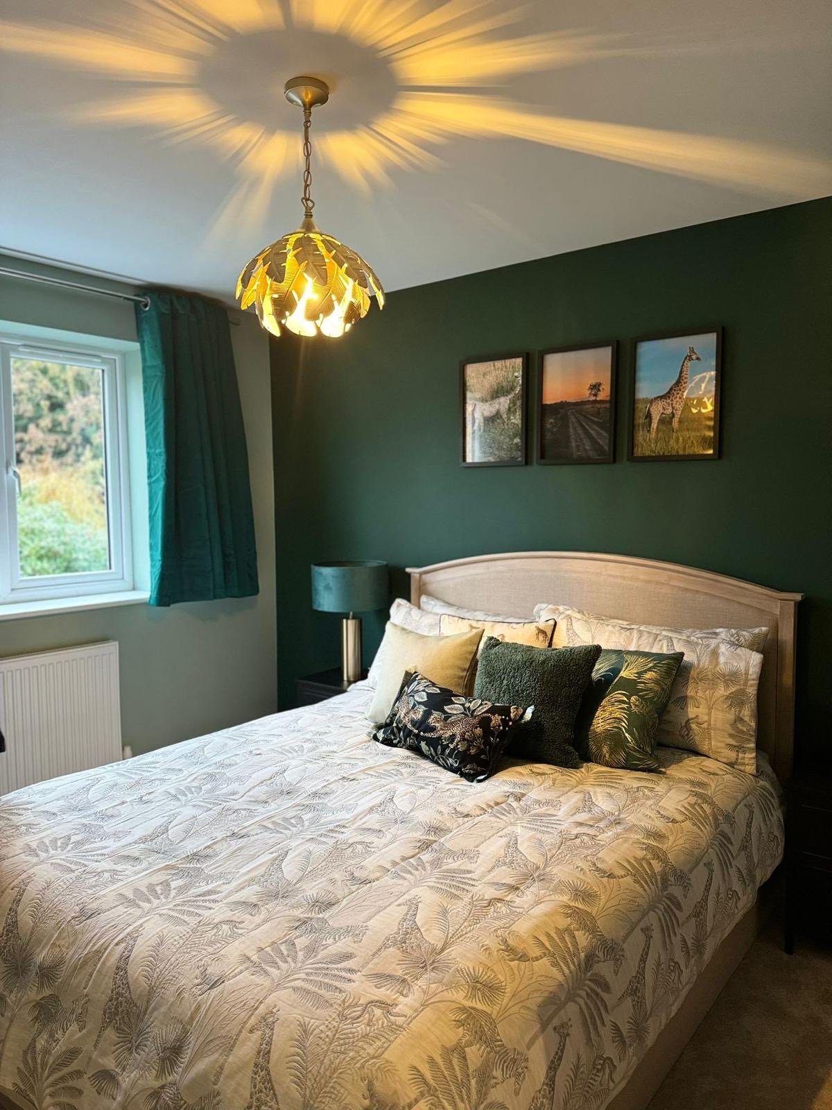 A bedroom with a bed with a patterned bedspread, multiple decorative pillows, a dark green accent wall, three framed pictures of nature scenes, a window with teal curtains, a bedside table with a teal lamp, and a ceiling light casting decorative shadows.