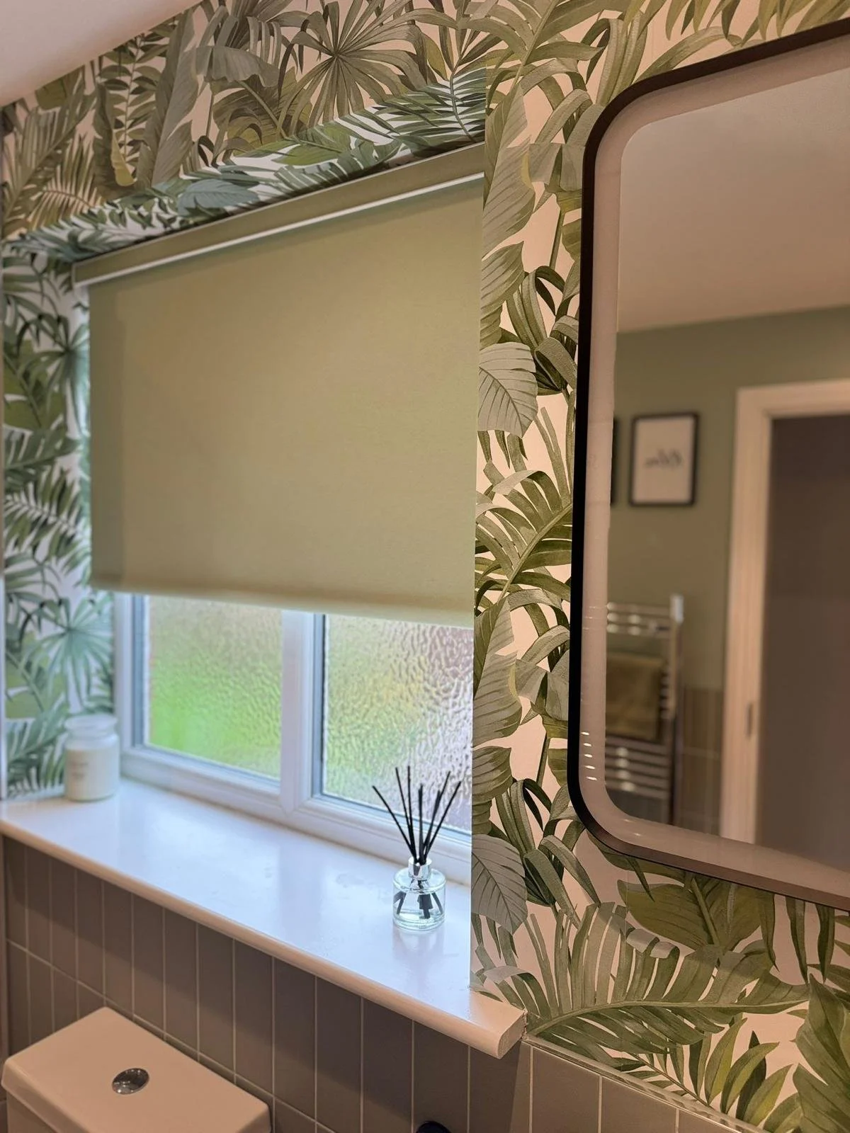 A bathroom with a green leafy wallpaper, a frosted window with a green roller blind, and a mirror with a black frame.