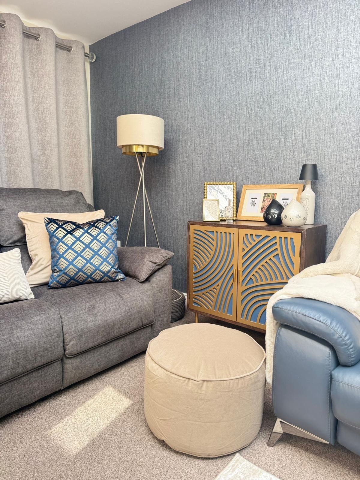 Living room with a gray sofa, blue armchair, beige ottoman, tall floor lamp, and a wooden cabinet with decorative vases and picture frames against a textured blue wall.