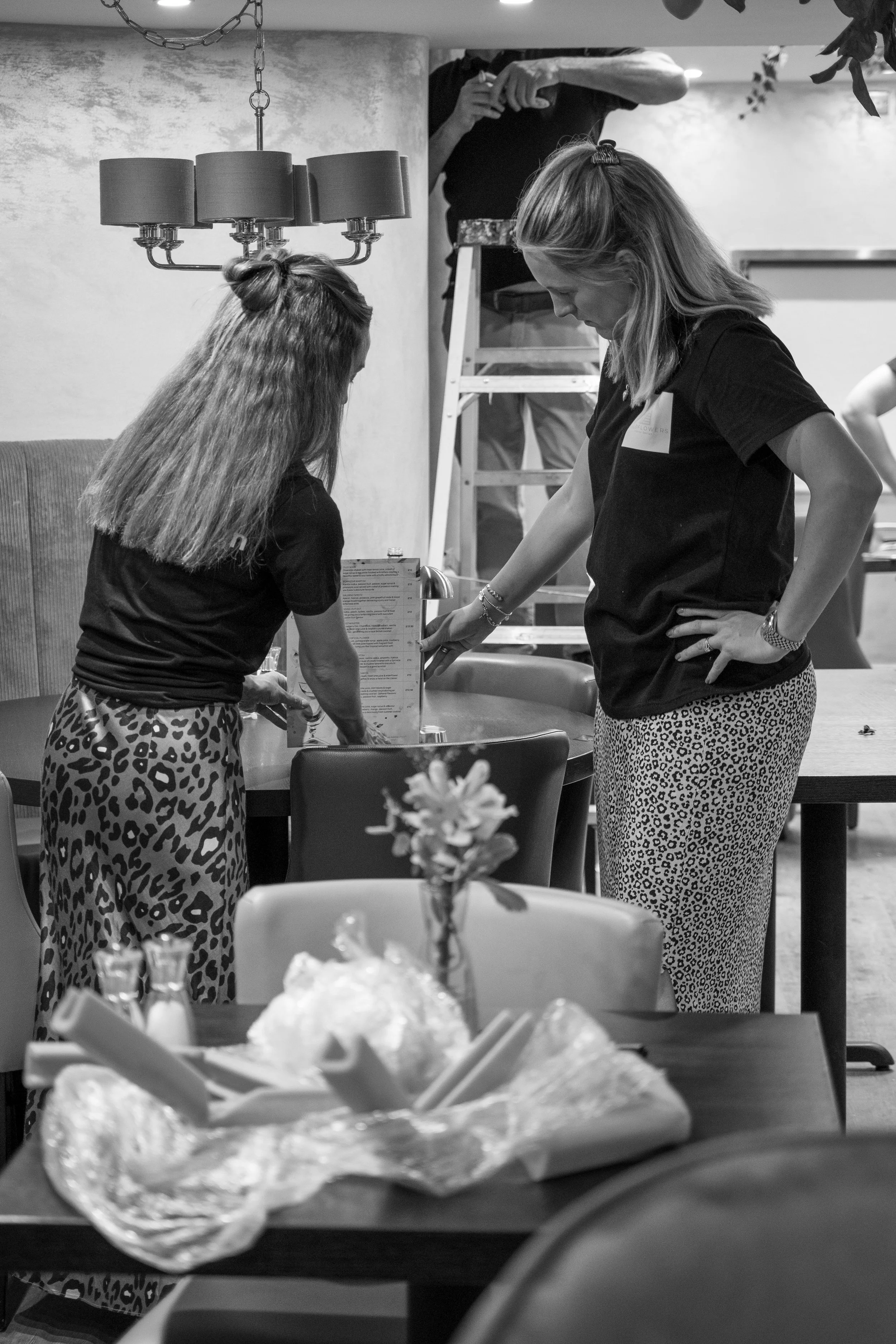 Two women standing at a table, looking at a menu, one with hand on hips, in a room with a chandelier and a flower centerpiece on another table.