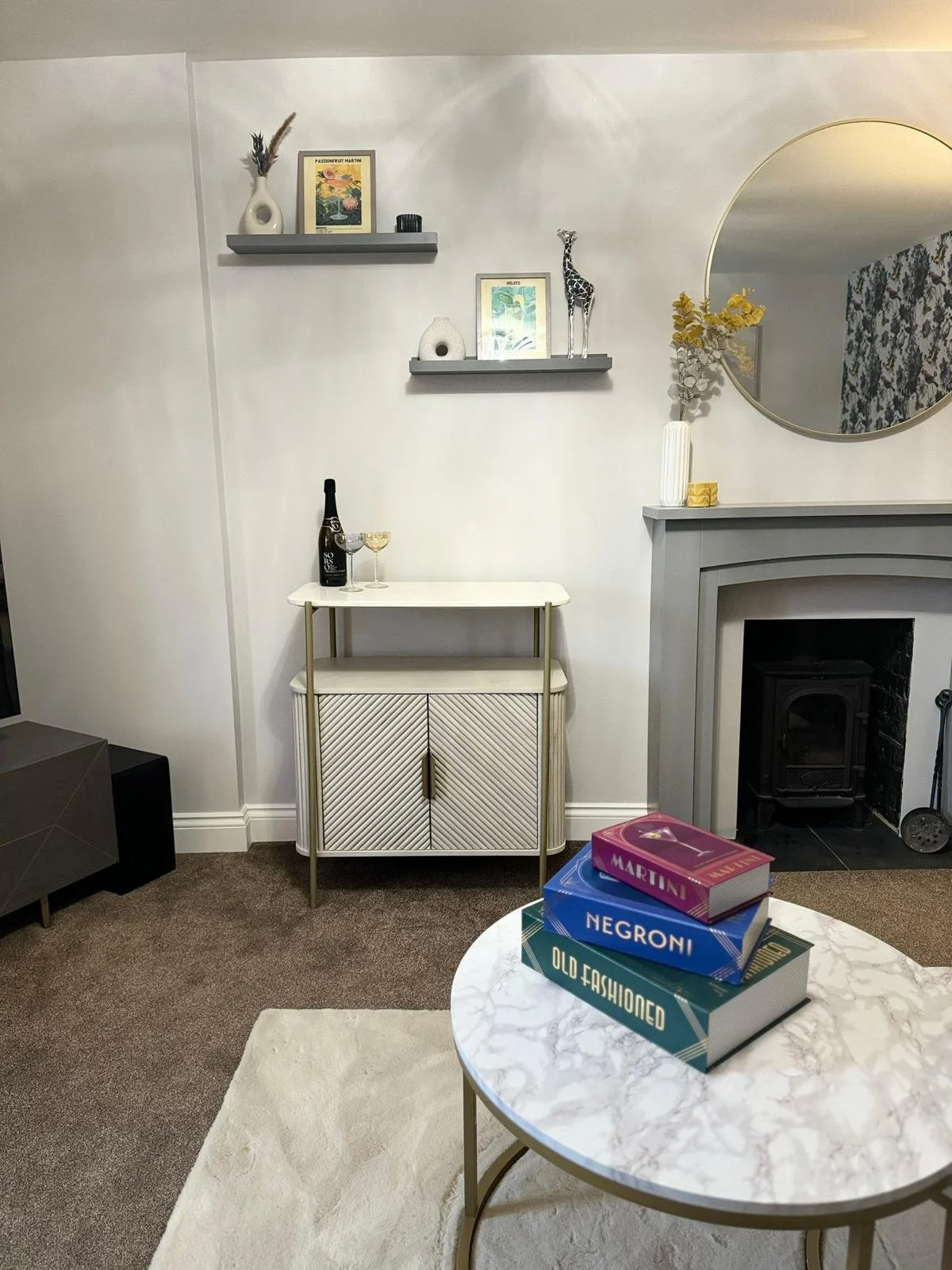 A living room with a white marble-topped coffee table with three colorful books stacked on it. Behind it is a light-colored cabinet with a champagne bottle and glass on top. To the right is a fireplace with a gray mantel and a round mirror above it, 