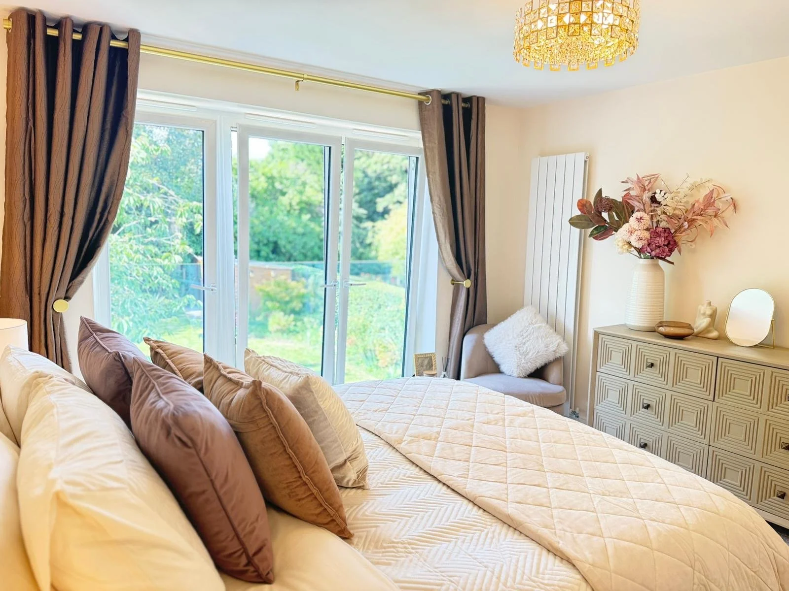 A cozy bedroom with a large bed filled with beige, brown, and purple pillows. Large windows with dark curtains reveal greenery outside. A dresser decorated with a vase of pink and white flowers, a mirror, and decorative objects. A chair with a furry 