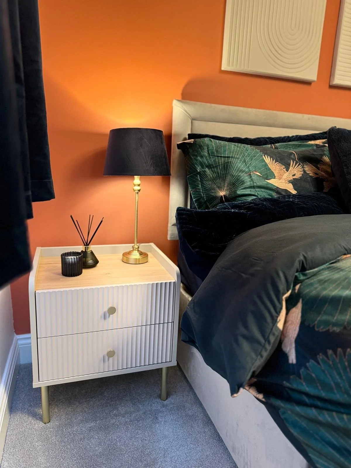 A bedside table with two drawers, a black lamp, and a container with reed diffusers. A bed with dark bedding and tropical leaf and bird patterned pillows is next to the table. The wall behind is painted a warm color.