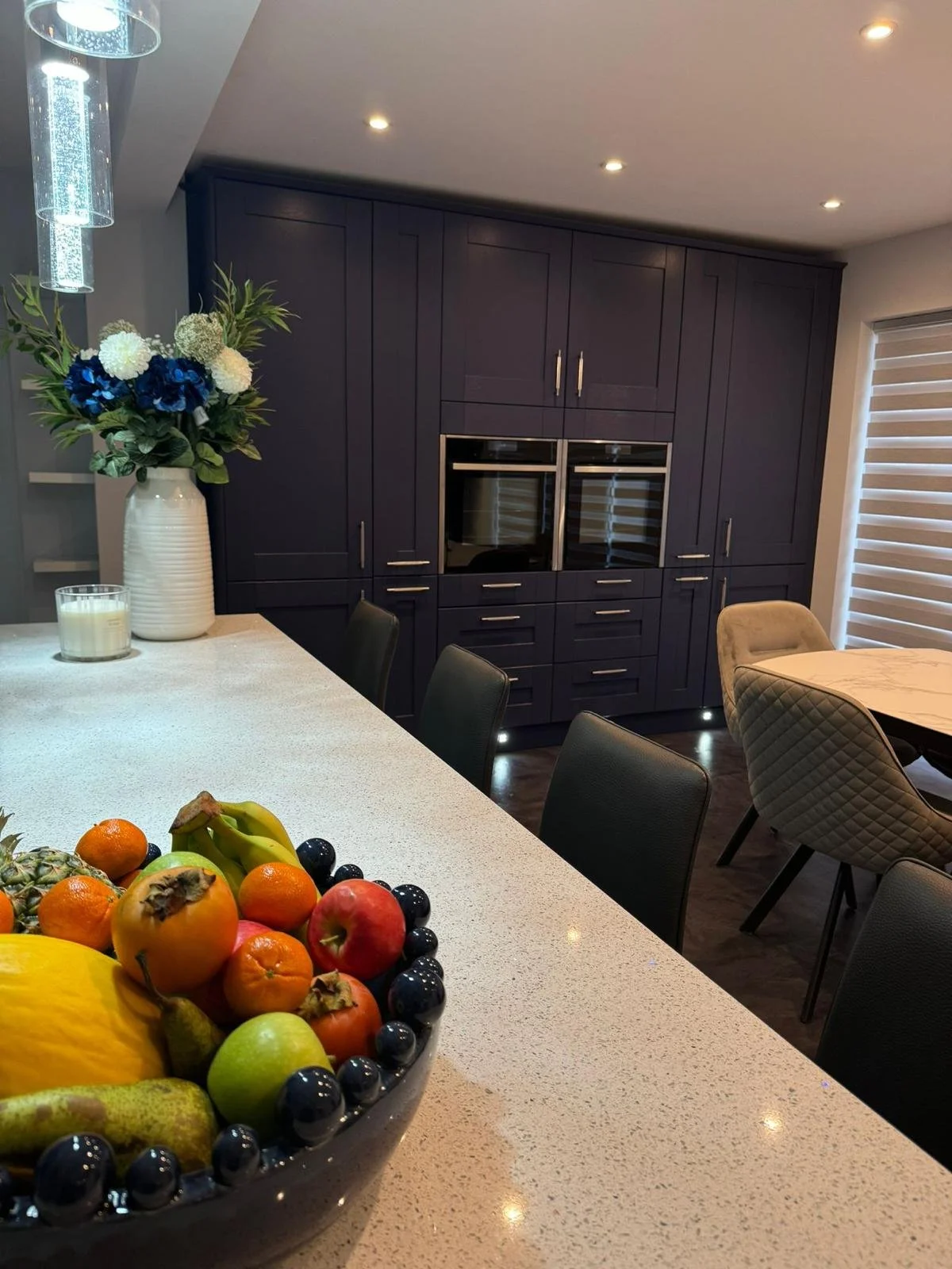 A kitchen dining area with a large fruit bowl on a white countertop, dark cabinetry with built-in appliances, a vase with flowers, and a window with blinds.