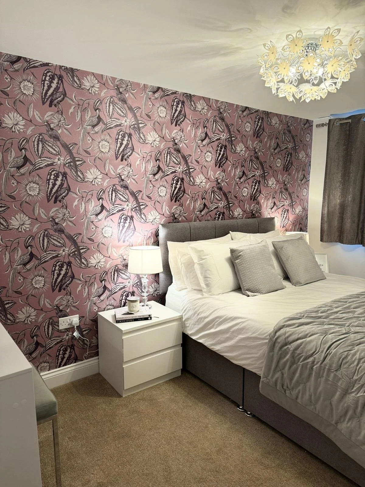 A bedroom with a floral-patterned pink wallpaper, a bed with white and gray pillows, and a gray headboard. There is a white nightstand with a lamp, a book, and a candle, and a chandelier hanging from the ceiling. A gray curtain covers a window.
