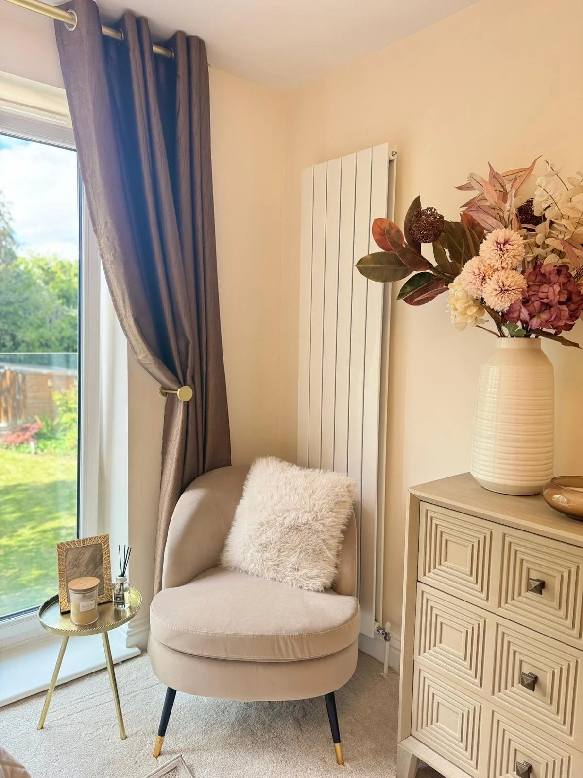 A cozy corner with a beige armchair with a fluffy white pillow, a small gold side table with a candle, a photo frame, and reed diffuser. There's a window with brown curtains, a cream-colored dresser, and a large vase with a floral arrangement of pink