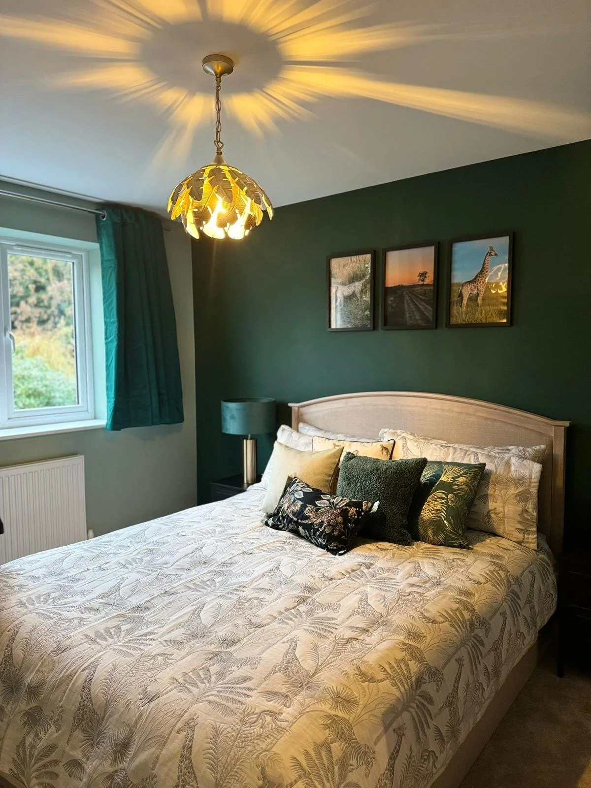 A bedroom with a bed, decorative pillows, a window with teal curtains, a dark green accent wall with three framed pictures, a bedside table with a teal lamp, and a ceiling light casting star-shaped shadows