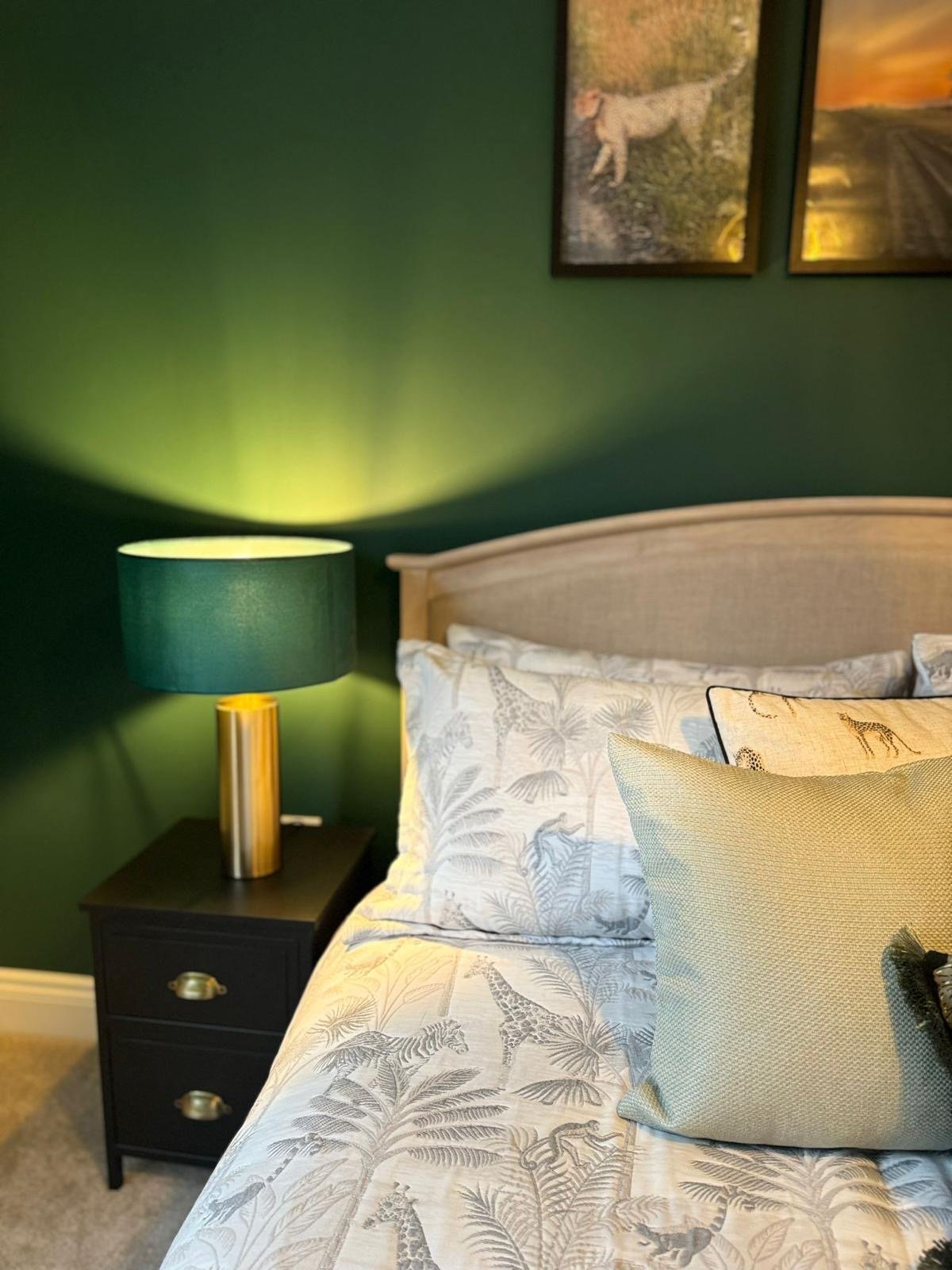 A bedroom with a beige headboard, a bed with pillowcases featuring animal and jungle prints, a beige decorative pillow, a black nightstand with two drawers and gold handles, a cylindrical table lamp with a green shade and gold base, green walls, and 