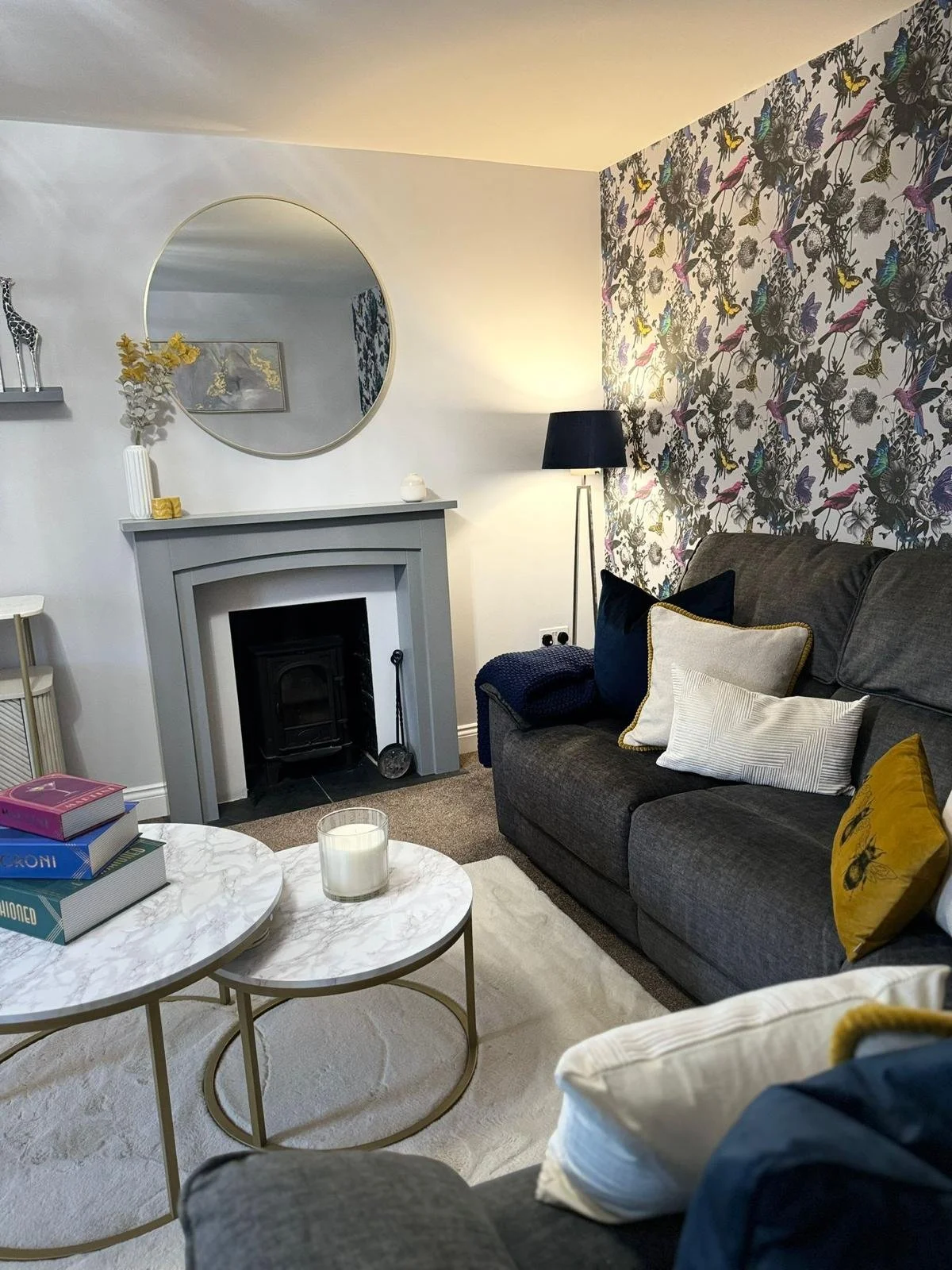Cozy living room with a gray sofa, pillows, a fireplace with a round mirror above it, a marble coffee table with books and a candle, a floral wallpaper accent wall, and a standing floor lamp.