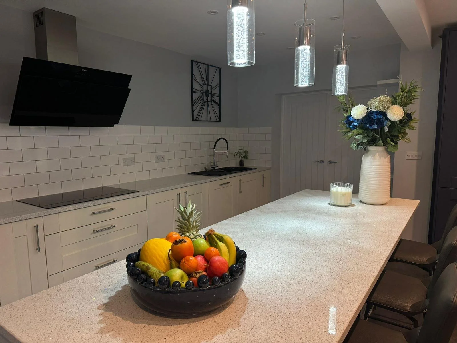 Modern kitchen with a white countertop, a black fruit bowl filled with various fruits, a white vase with blue and white flowers, a candle, and hanging pendant lights.